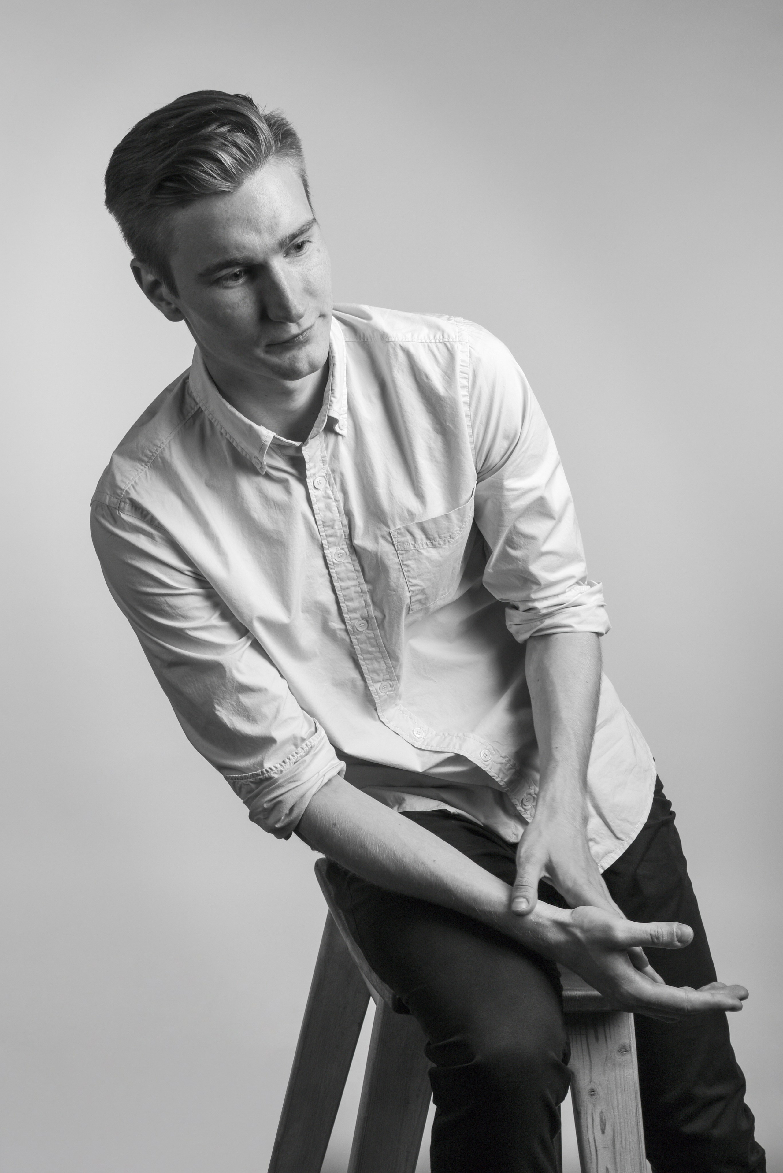 Фотосъёмка в студии, молодой человек на высоком табурете, сильно наклонился в сторону. Кажется, что теряет равновесие.