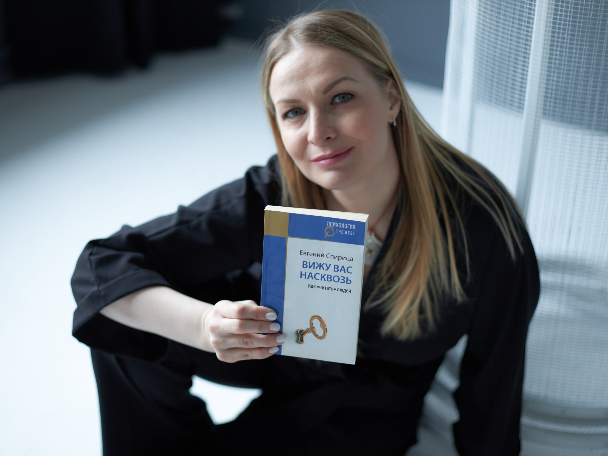 Блондинка с длинными волосами и в черном кимоно сидит на полу и держит перед собой книгу