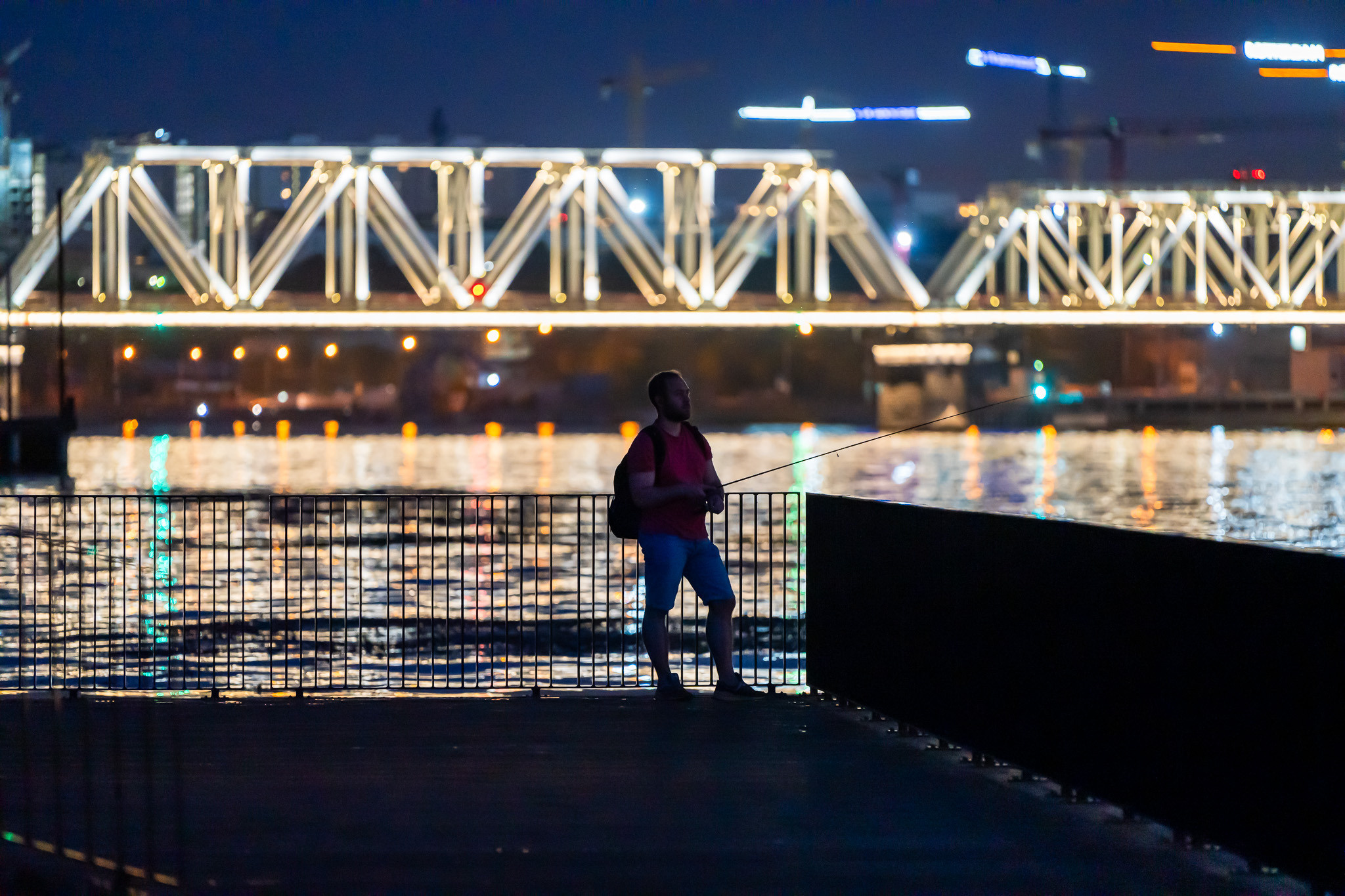 Рыбак на набережной Марка Шагала ночью на фоне светящегося моста