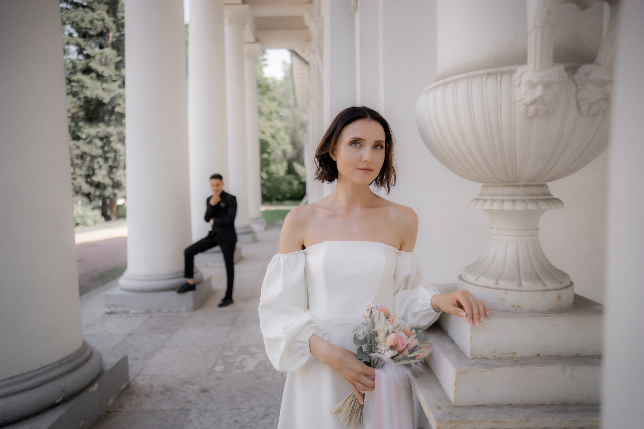 передний план - невеста в платье с открытыми плечами, задний план - жених в черном костюме на елагином острове