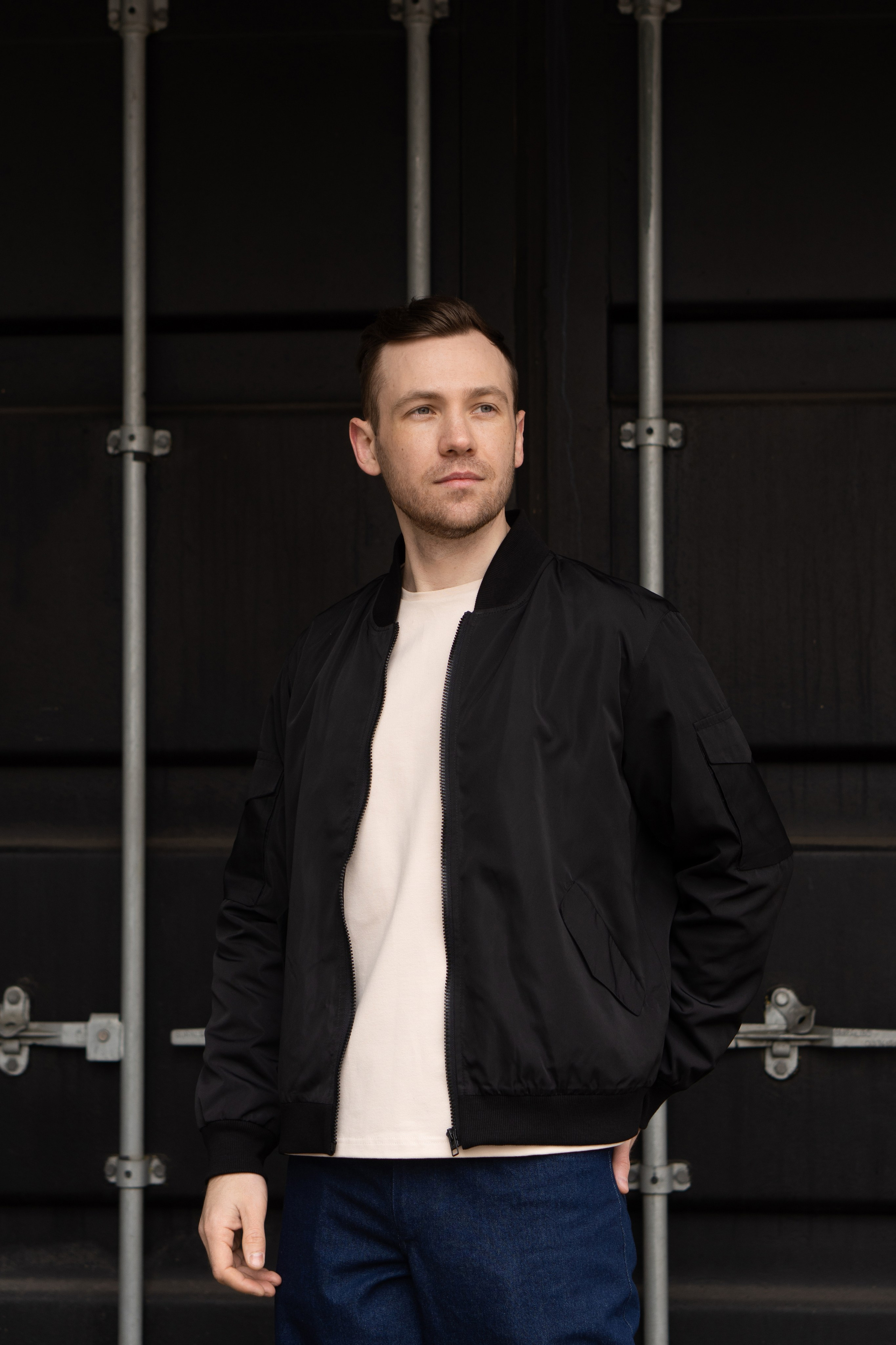 Мужчина с короткой щетиной в черном бомбере и синих джинсах стоит на фоне черного контейнера