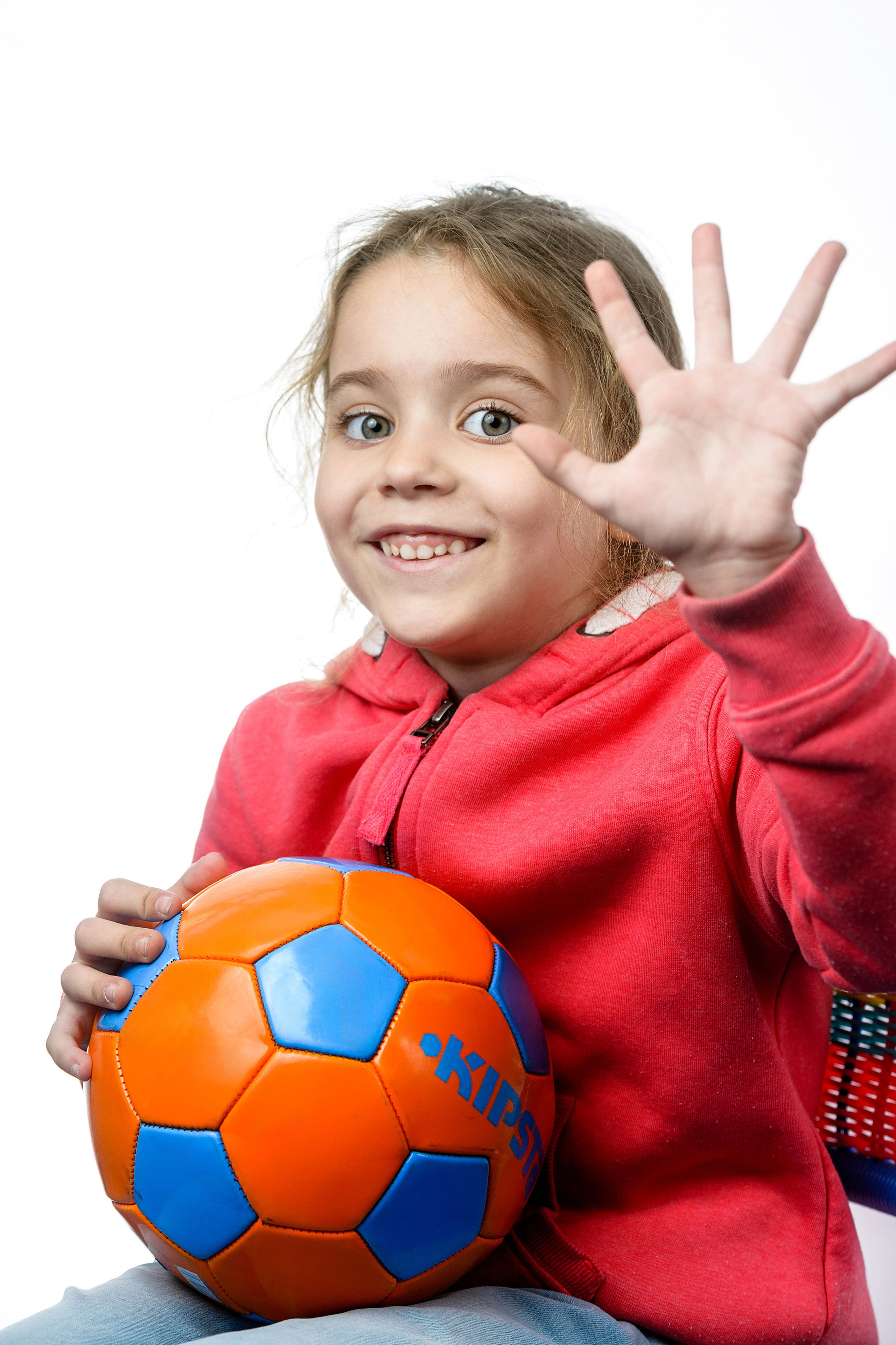 Девочка в красной толстовке держит синий мяч и показывает ладонь в студийной съёмке на белом фоне.