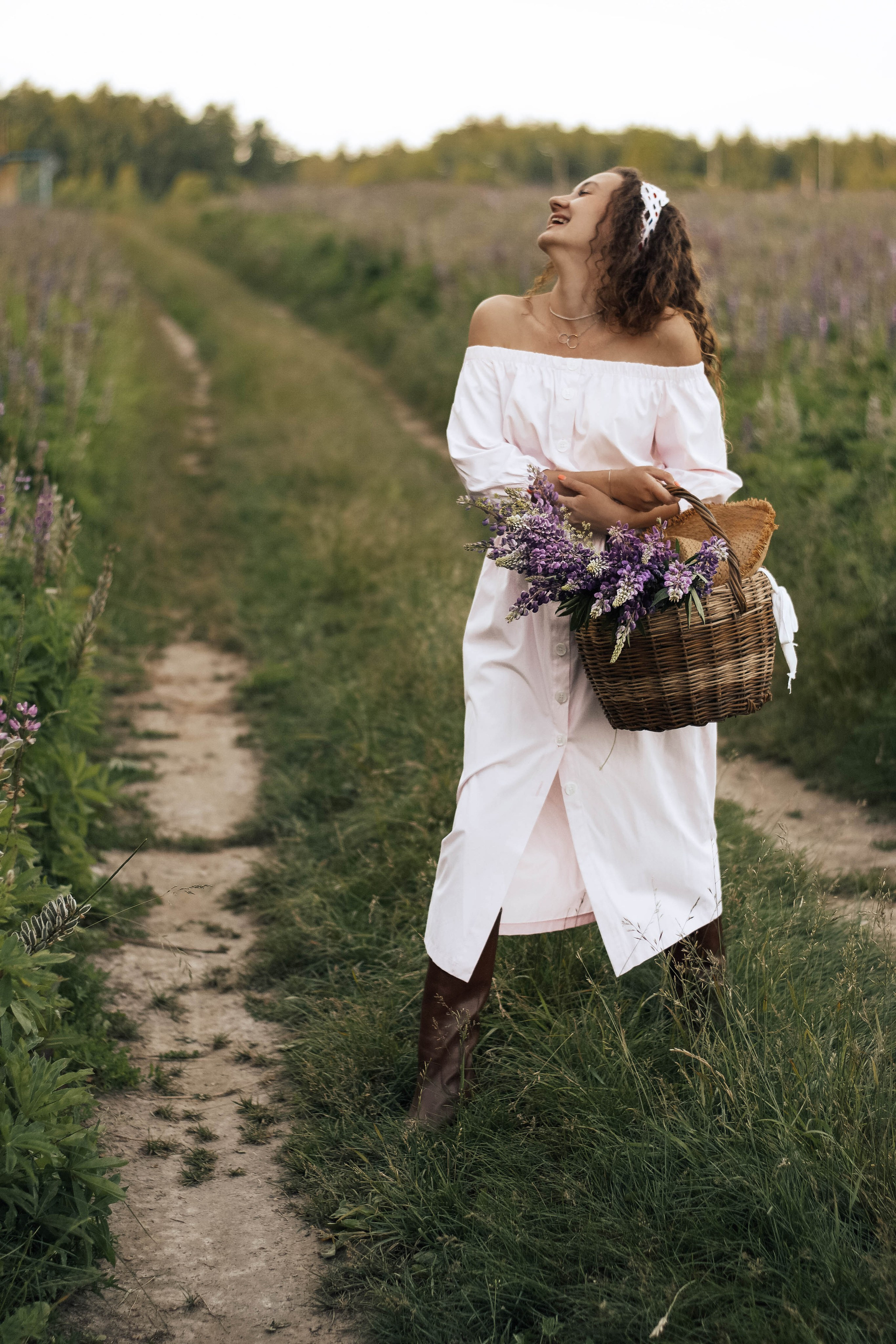 Девушка в белом платье идет по тропинке среди лавандовых полей, держа корзинку с цветами.