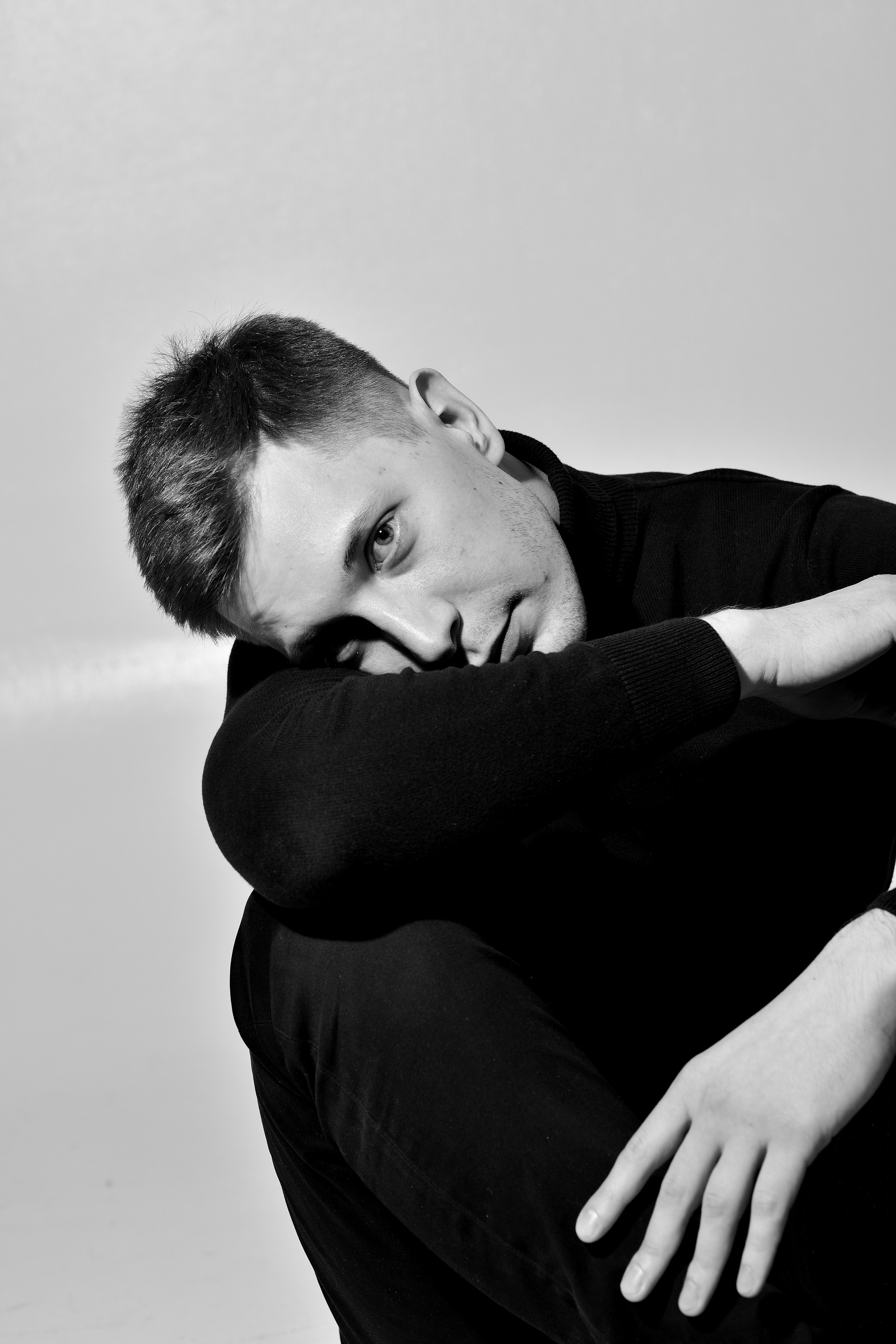 Портрет юноши в черной водолазке на фоне белой стены.