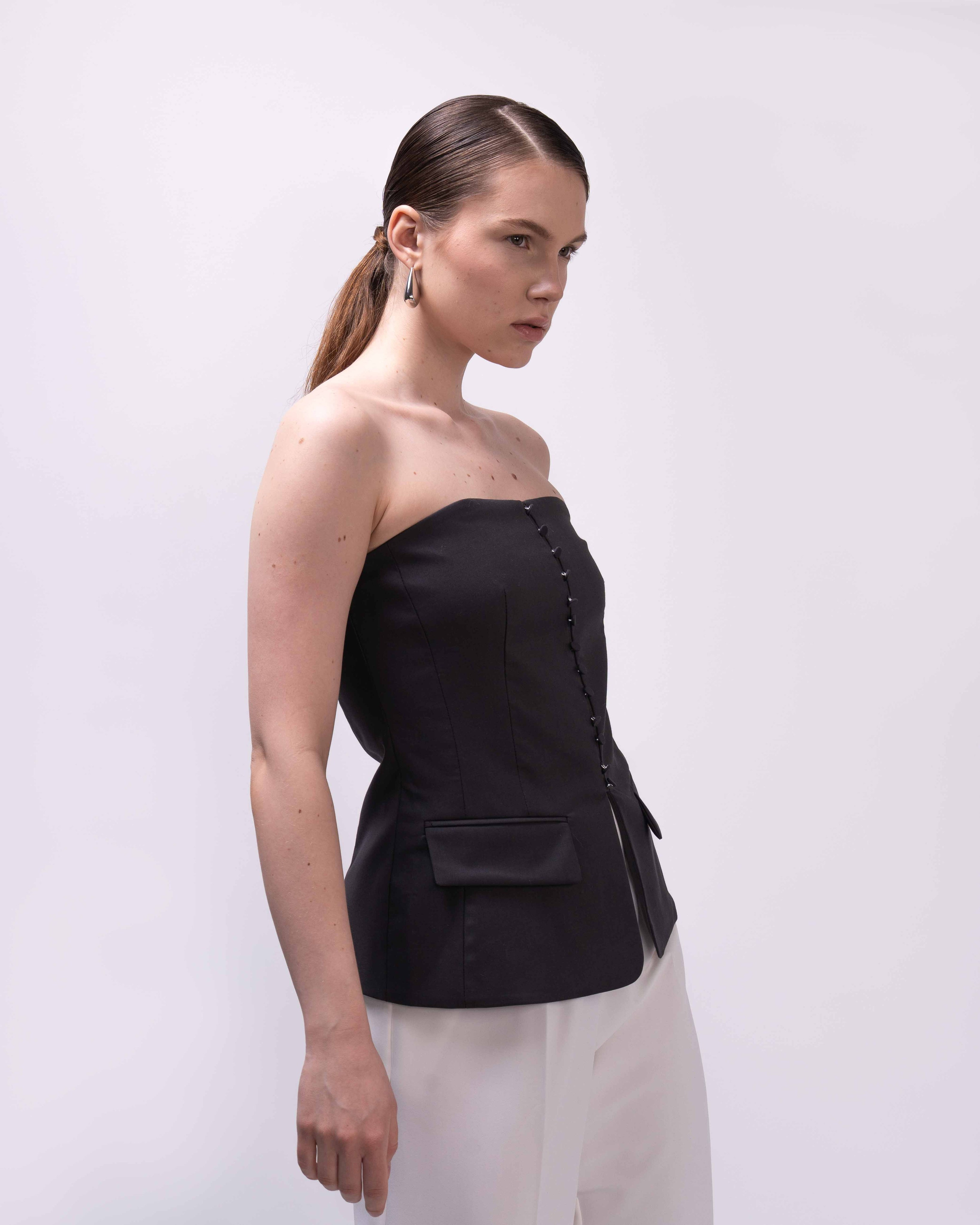 Фото товара для каталога  Ozon или Wildberries.  На фотографии изображена женщина, которая стоит на фоне светлой стены. Она одета в черный топ без рукавов, который имеет декоративные элементы, такие как шнуровка и завязки. Брюки у нее белые, что создает контраст с верхней частью одежды. Волосы женщины собраны в хвост, а на ухе виден серьга. Её взгляд направлен в сторону, что придает изображению определенную динамику и глубину.