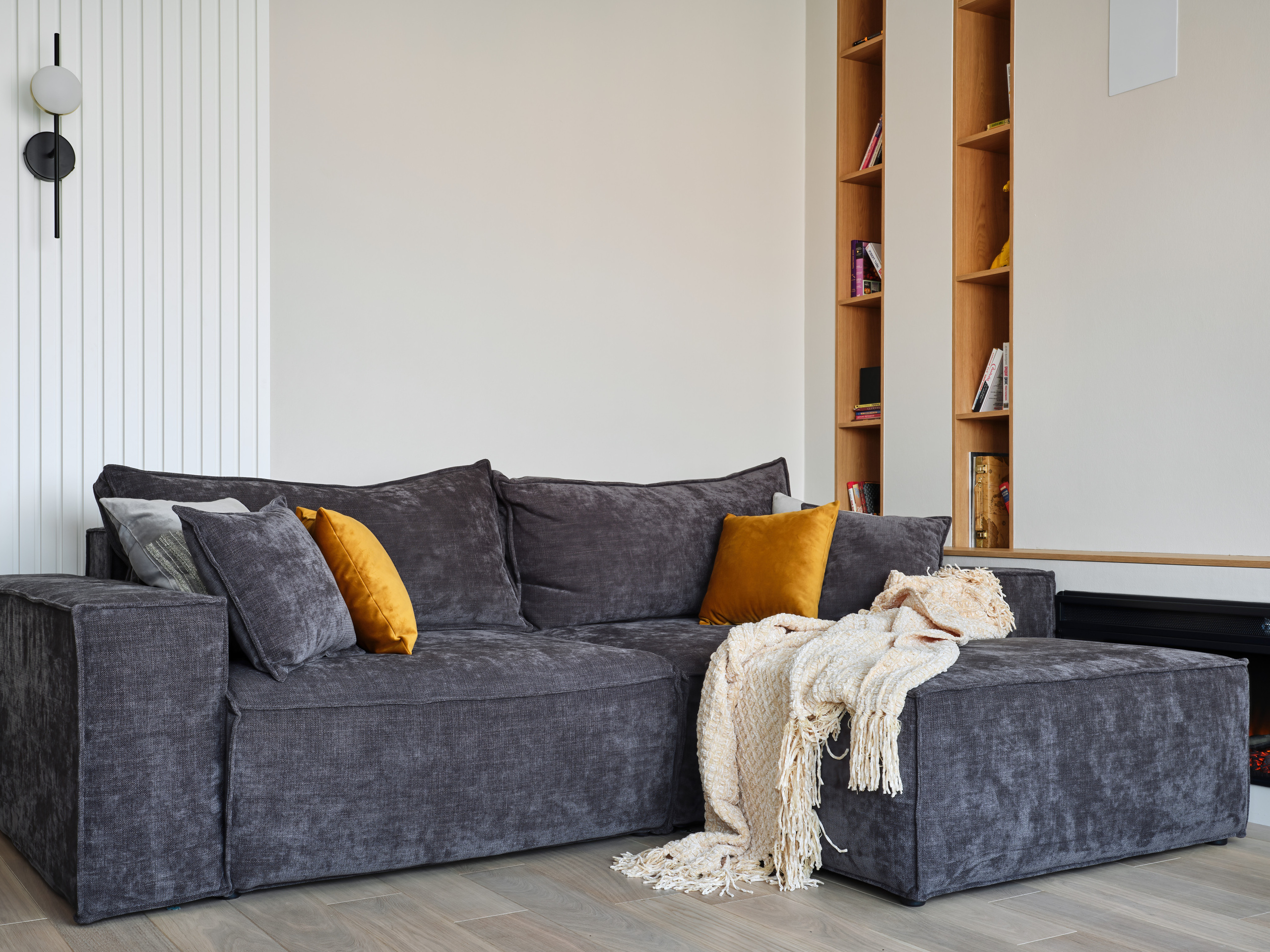 Гостиная в современном стиле с большим серым диваном, яркими горчичными подушками, встроенным стеллажом из дерева и мягким пледом. Дизайн интерьера разработан Анжеликой Исаевой, фотосъемка Сергея Денисенко.