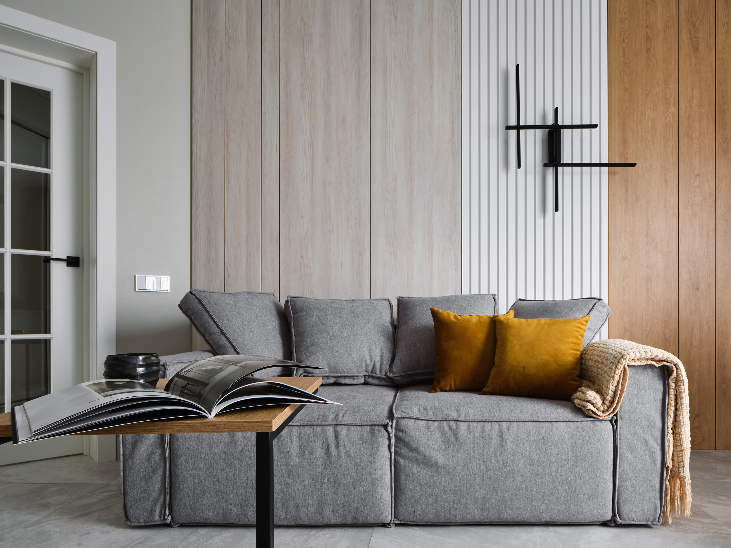 Уютная гостиная с серым диваном, декоративными подушками и деревянными панелями на стене. Проект дизайнера Анжелики Исаевой, интерьерная съемка Сергея Денисенко, современный стиль с акцентом на фактуры и теплую палитру.