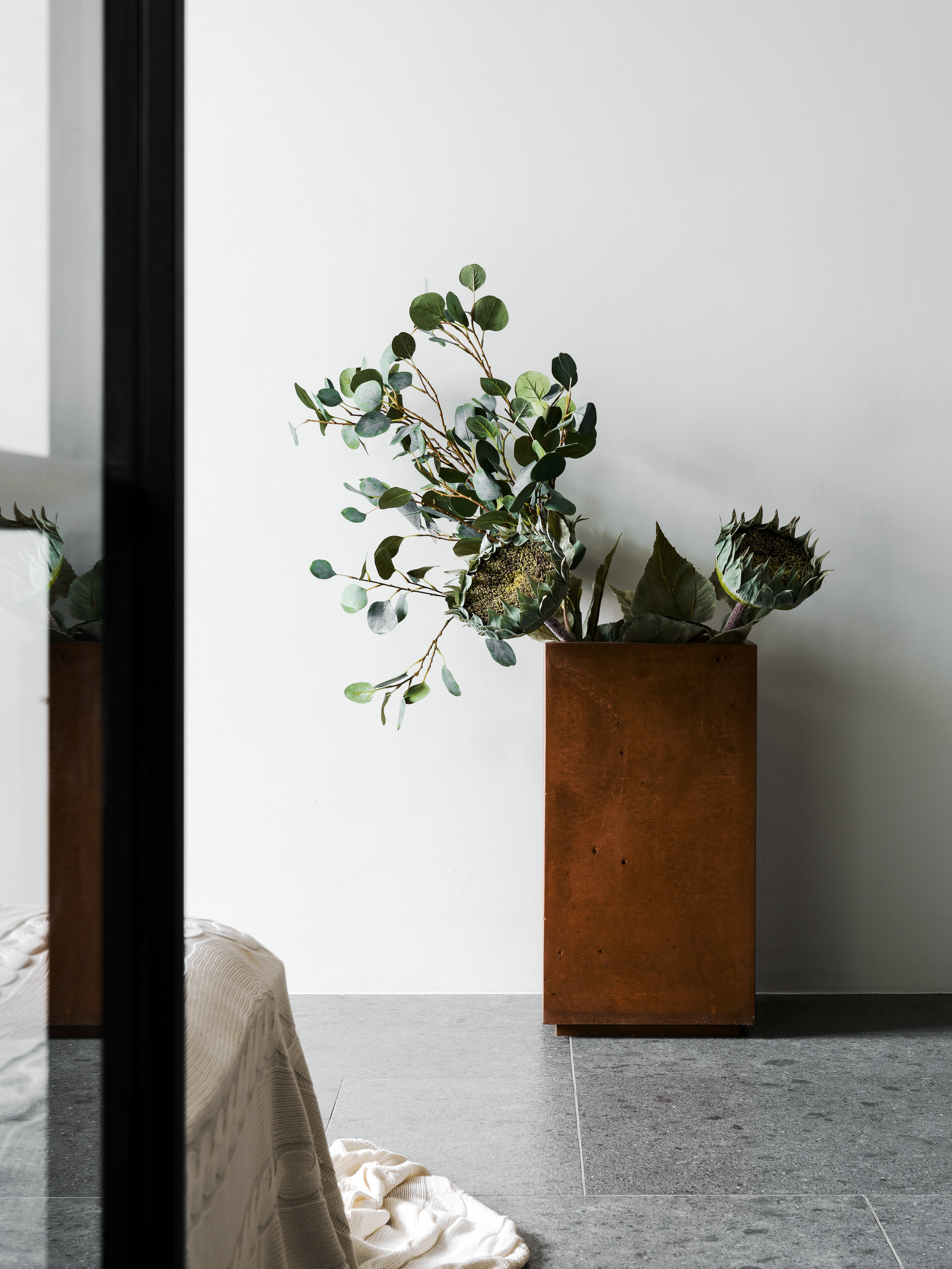 Декоративная деталь интерьера: стильный высокий кашпо с зелёным растением на фоне белой стены и серого каменного пола. Авторский минимализм дизайнеров Андрея Нелень и Екатерины Проскуряковой, фотосъёмка Сергей Денисенко.