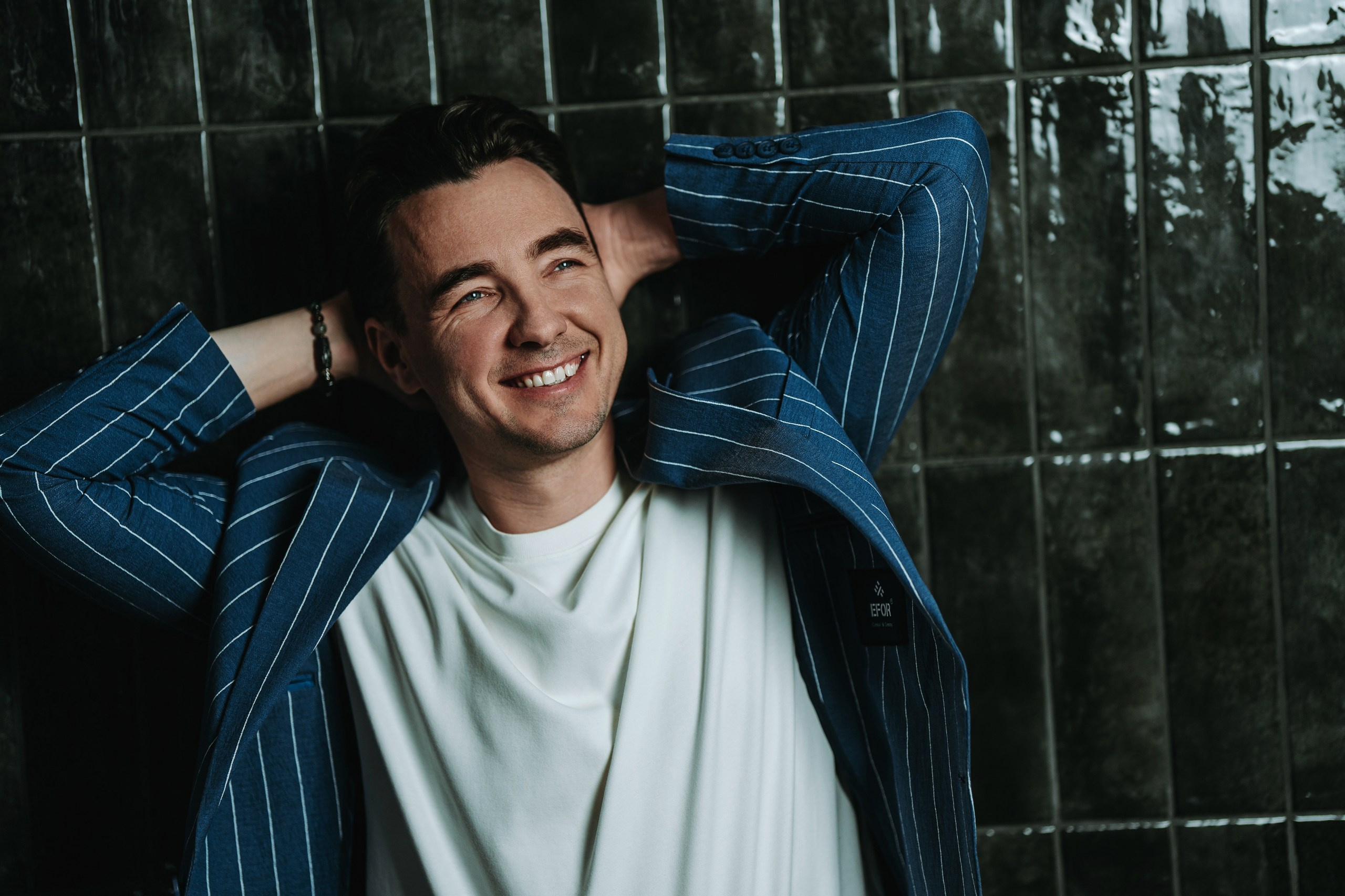 Улыбающийся мужчина в белой футболке и полосатом пиджаке запрокинул руки за голову на фоне тёмной глянцевой плитки.