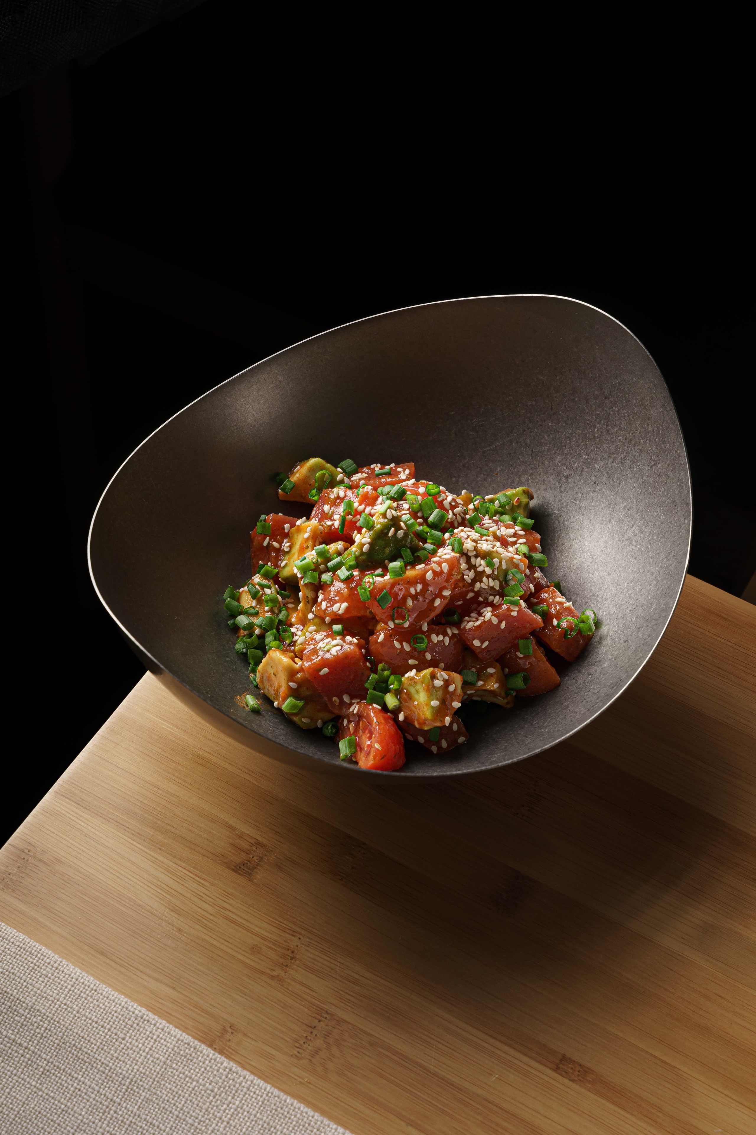 Poke bowl tuna with avocado, green onions, and sesame seeds, served in a matte black bowl on a wooden table. Top view.