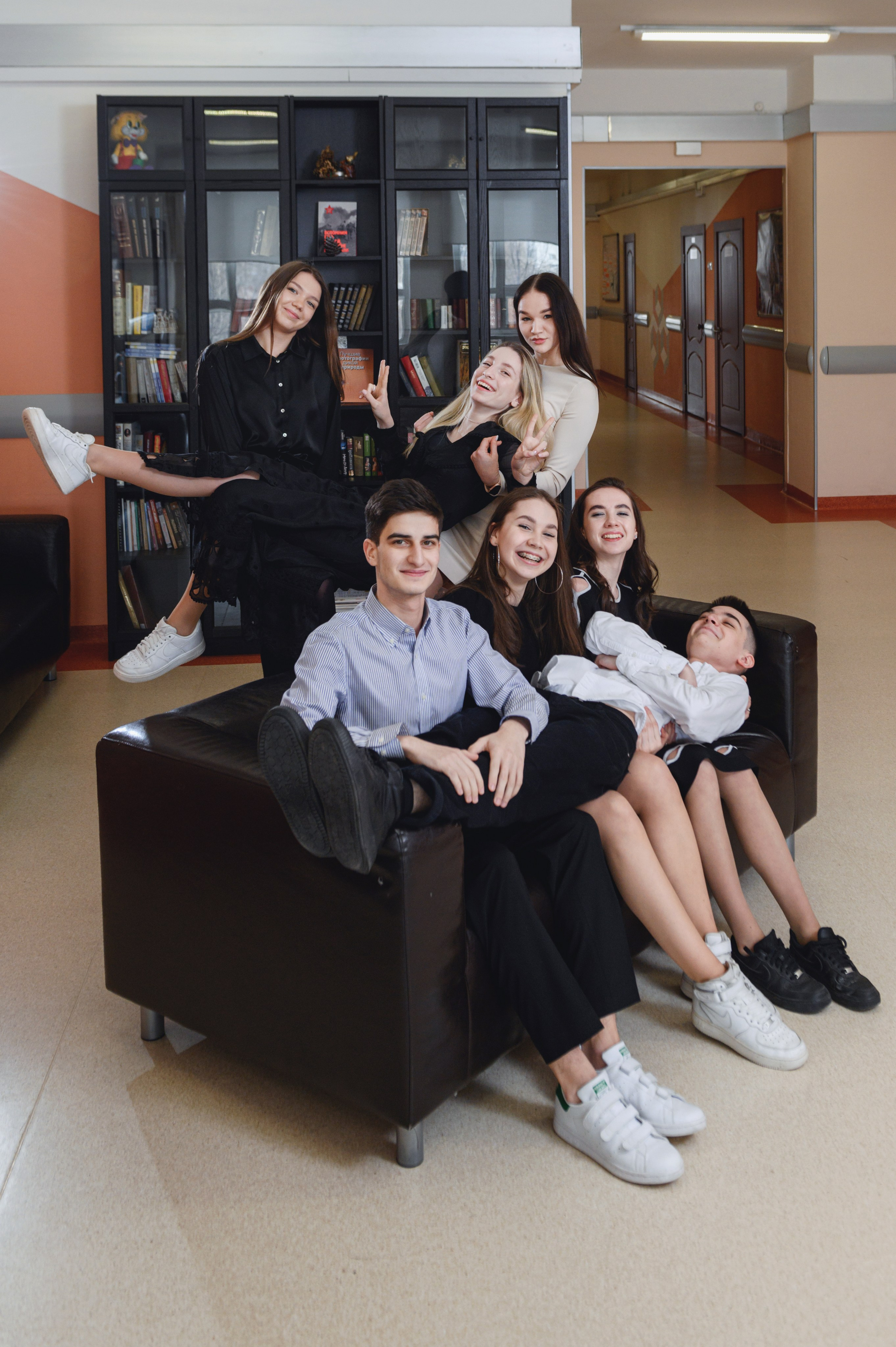 Группа старшеклассников разместилась на диване и рядом с ним в интерьерной локации для дружеского кадра.
