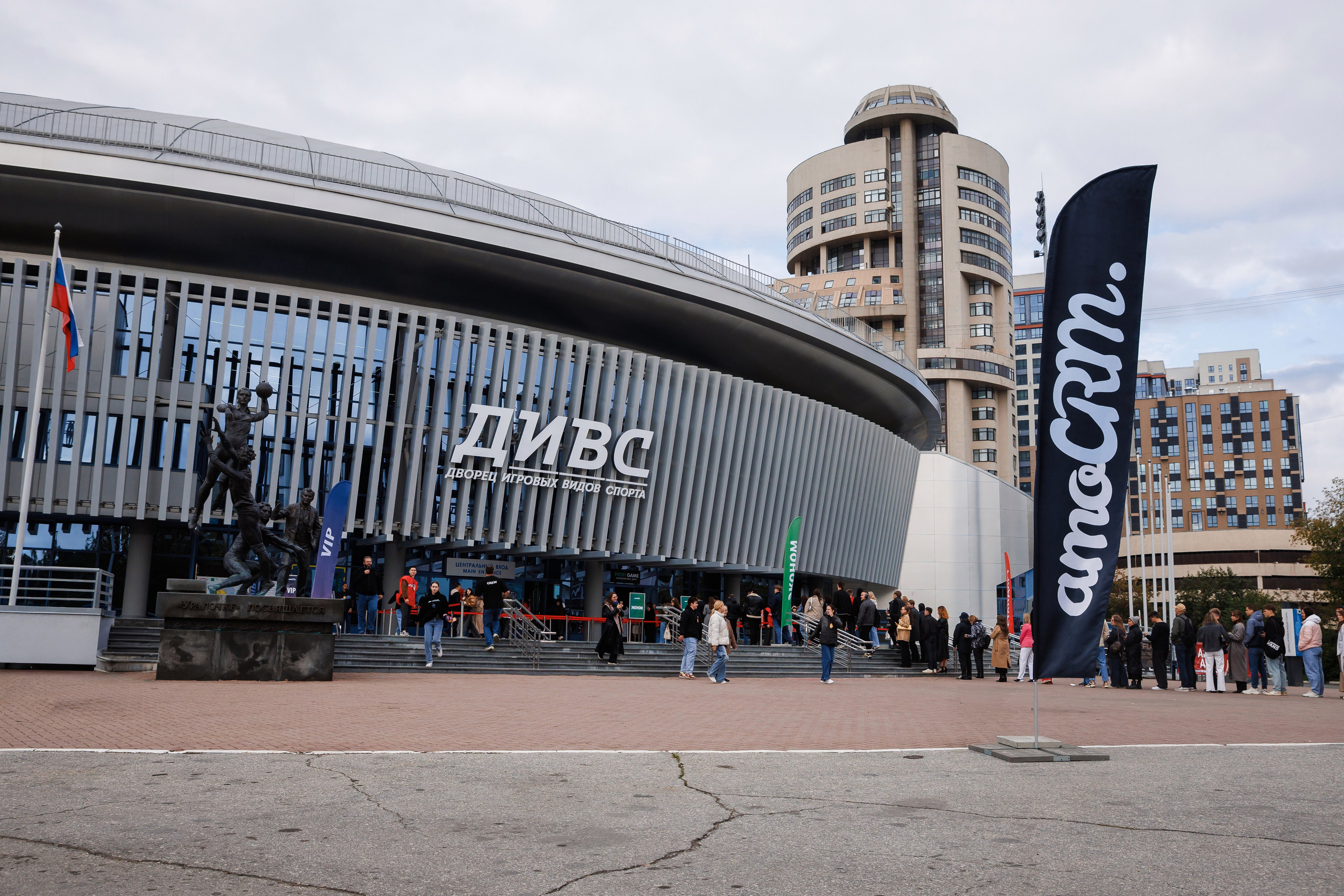 Люди стоят перед современной круглой ареной с вывеской «ДИВС» над входом. Рядом с входом развевается высокий черный флаг с надписью «amoCRM». На заднем плане видны высотные здания.