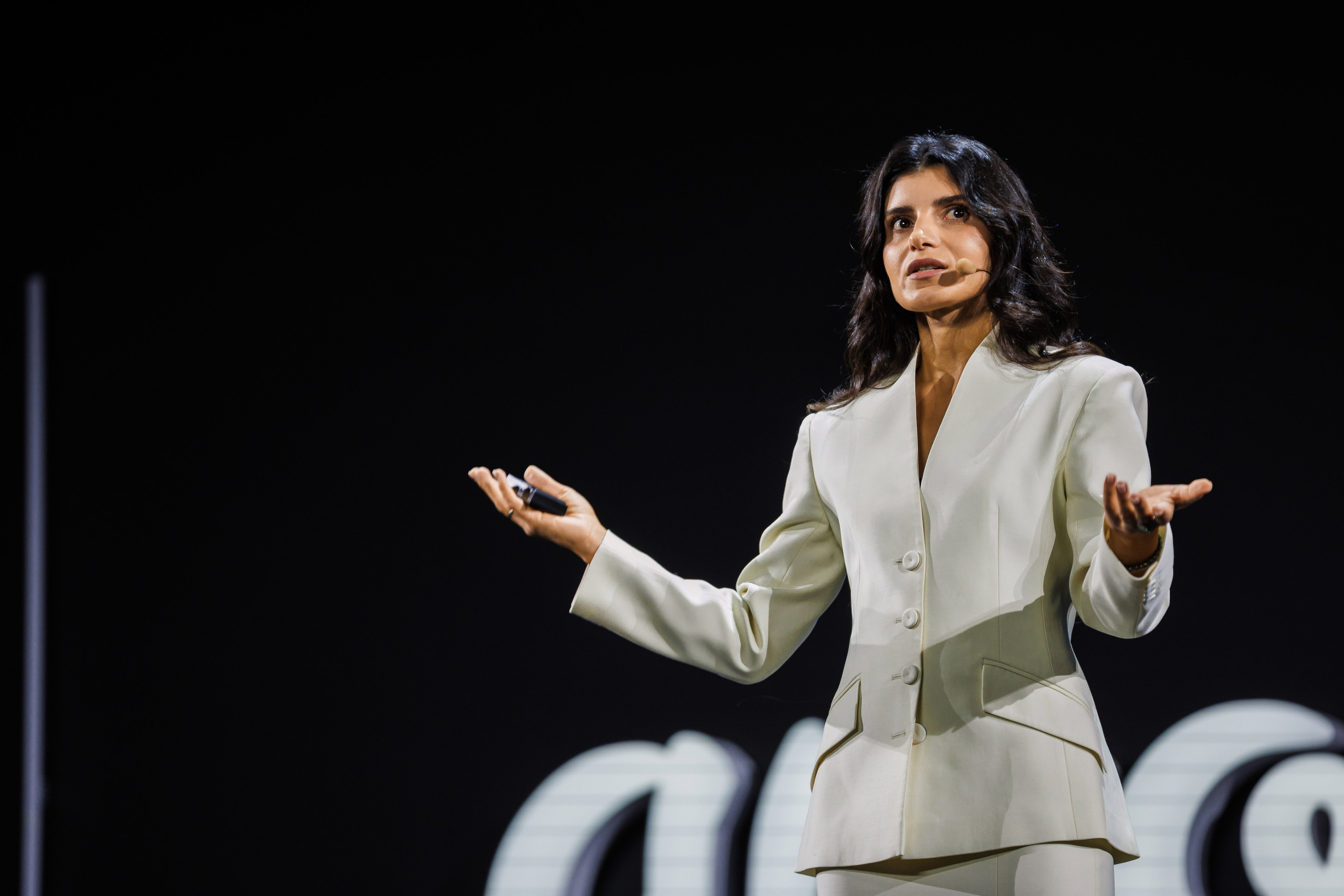 На сцене стоит женщина в светлом костюме с микрофоном в наушниках, жестикулируя руками во время выступления. За ней виден темный фон с частично белым текстом.