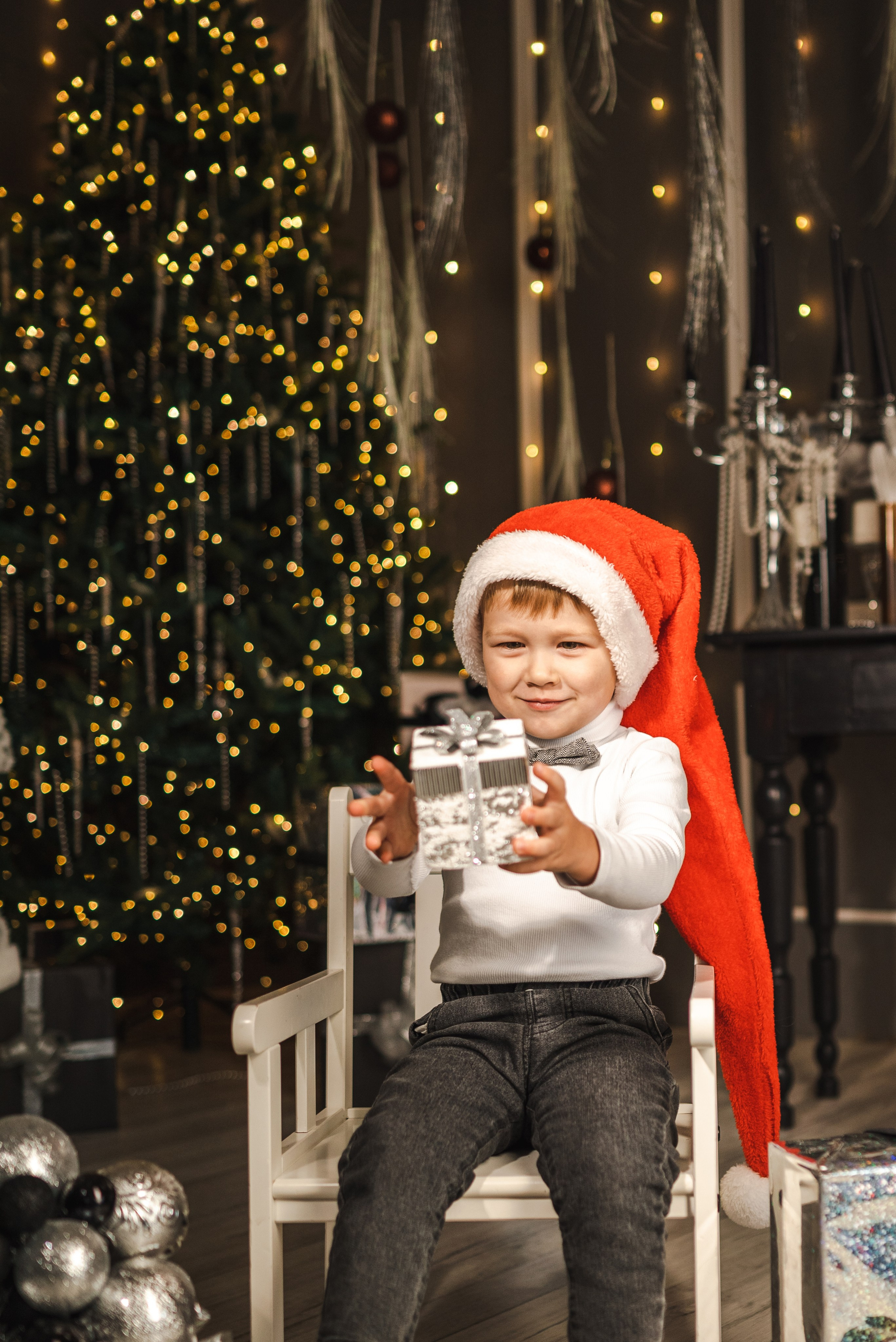 Детская семейная новогодняя фотосессия 2025 в фотостудии в Каменске-Шахтинском и Ростове-на-Дону. Ребенок стоит  с подарком на фоне новогодней елки. Фотограф Ольга Сагал 