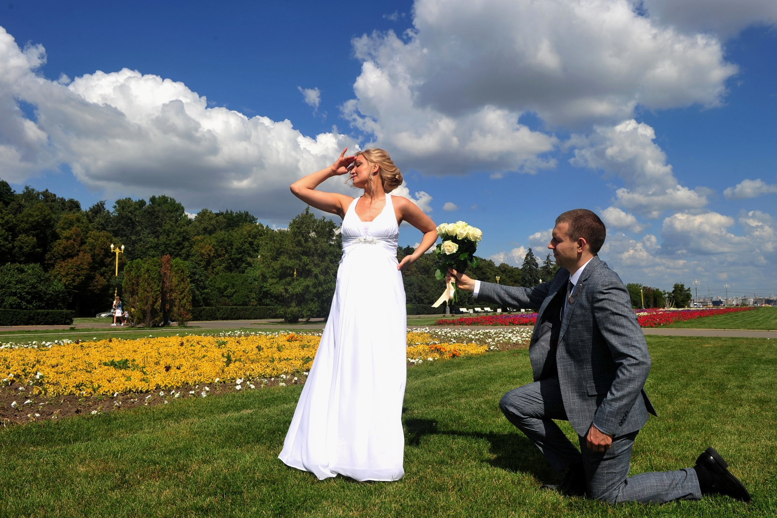 Свадьба на Воробьёвых Горах рядом с МГУ, жених стоит на одном колене и дарит свадебный букет невеста, невеста отвернулась, голубое небо, желтые цветы на клумбе, изумрудная зелень травы на лужайке