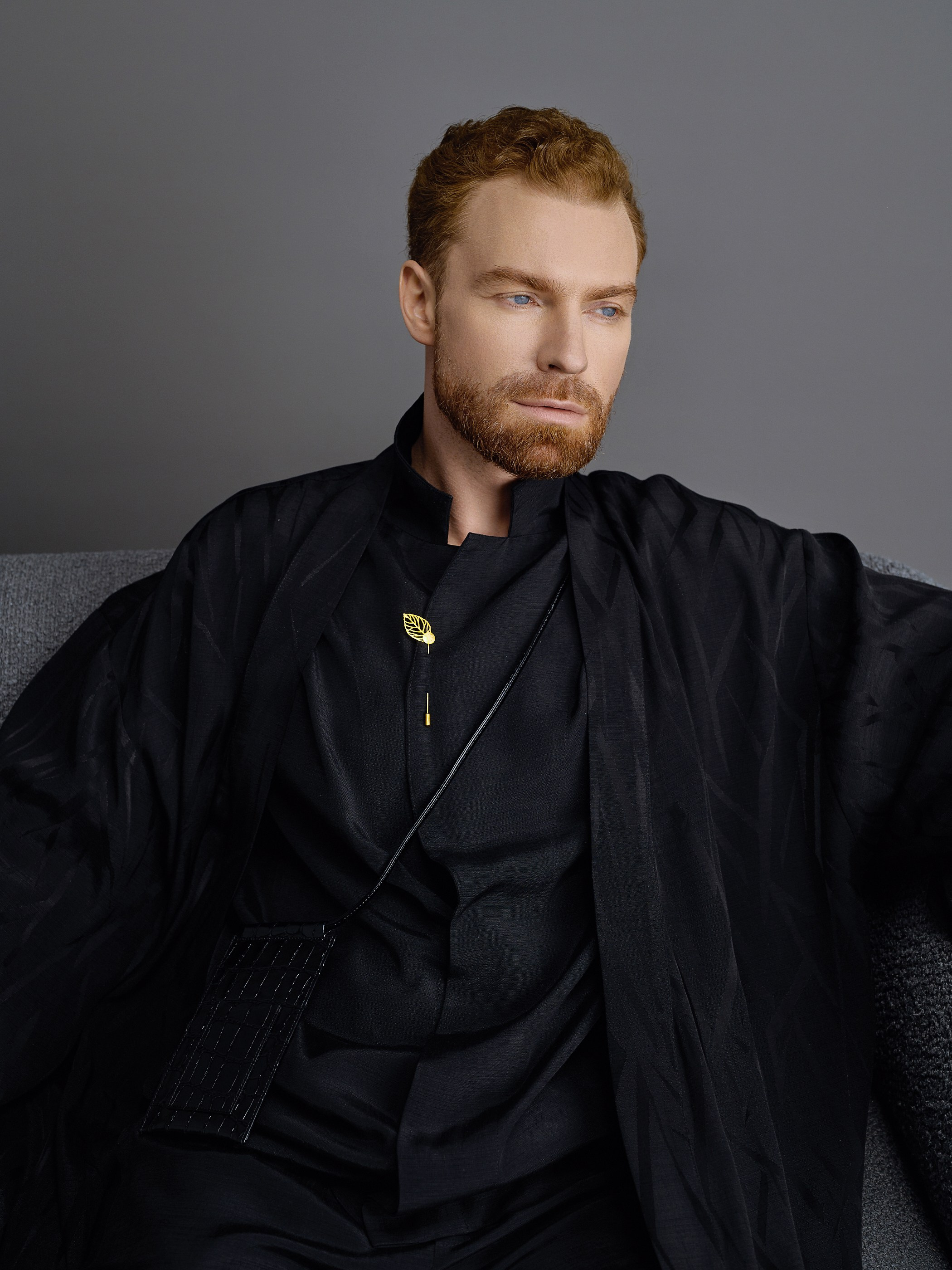 Портрет рыжеволосого мужчины в чёрной одежде с золотым аксессуаром на фоне дивана.