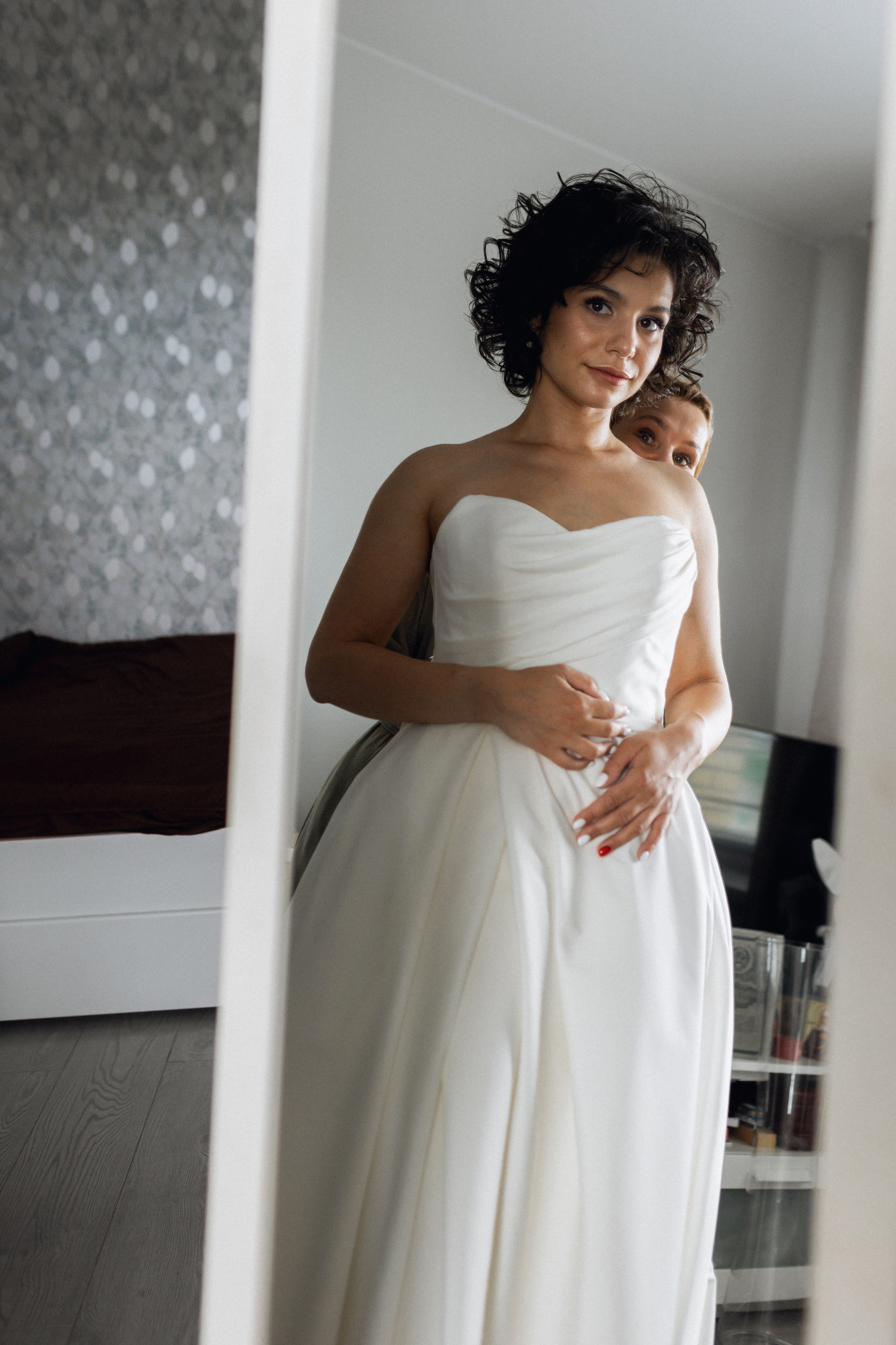 Эмоциональный кадр для салона NATALIE L: мама завязывает шнуровку на атласном платье невесты, а в зеркале отражается их общая улыбка 