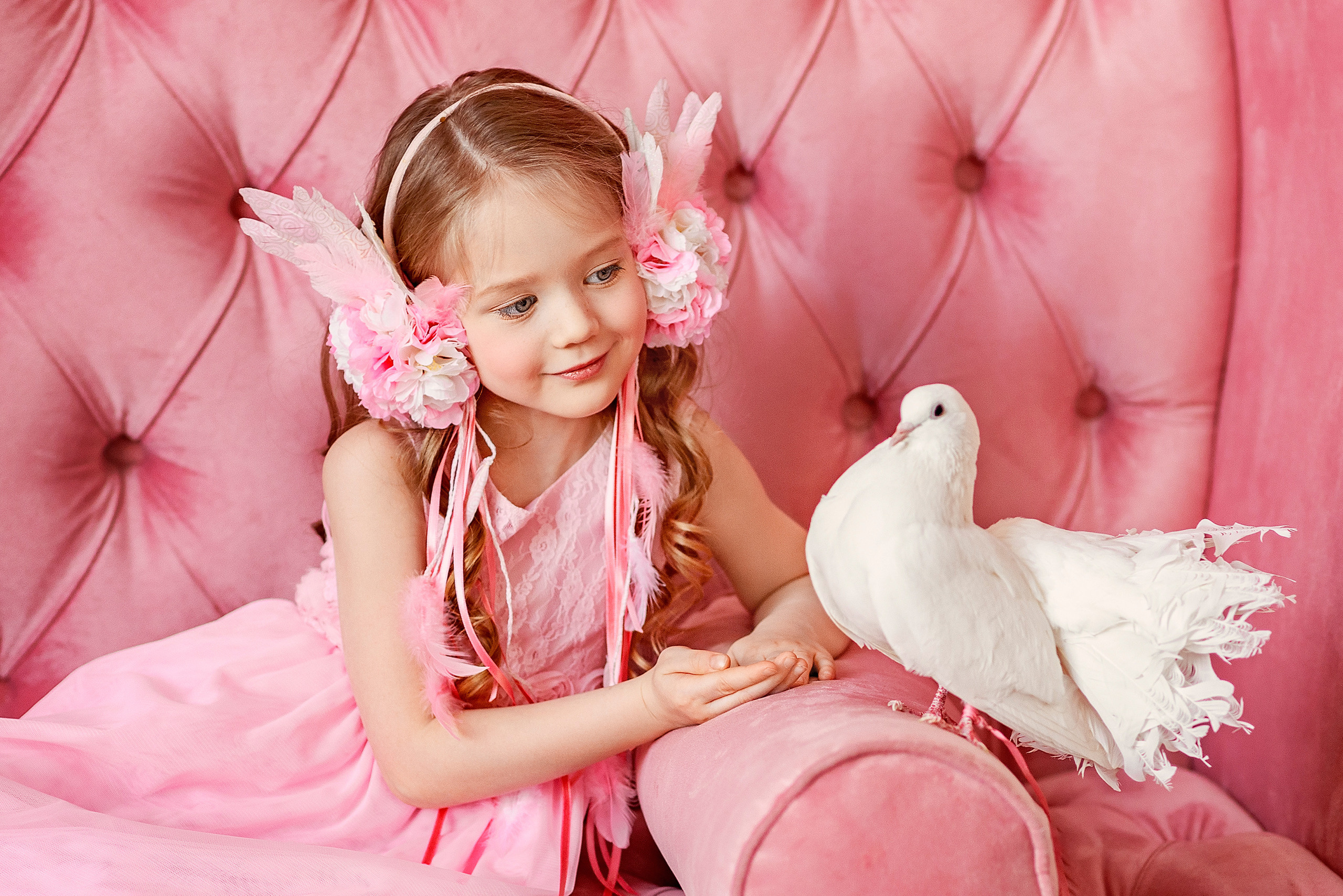 Девочка сидит на розовом диване и смотрит на белого голубя.