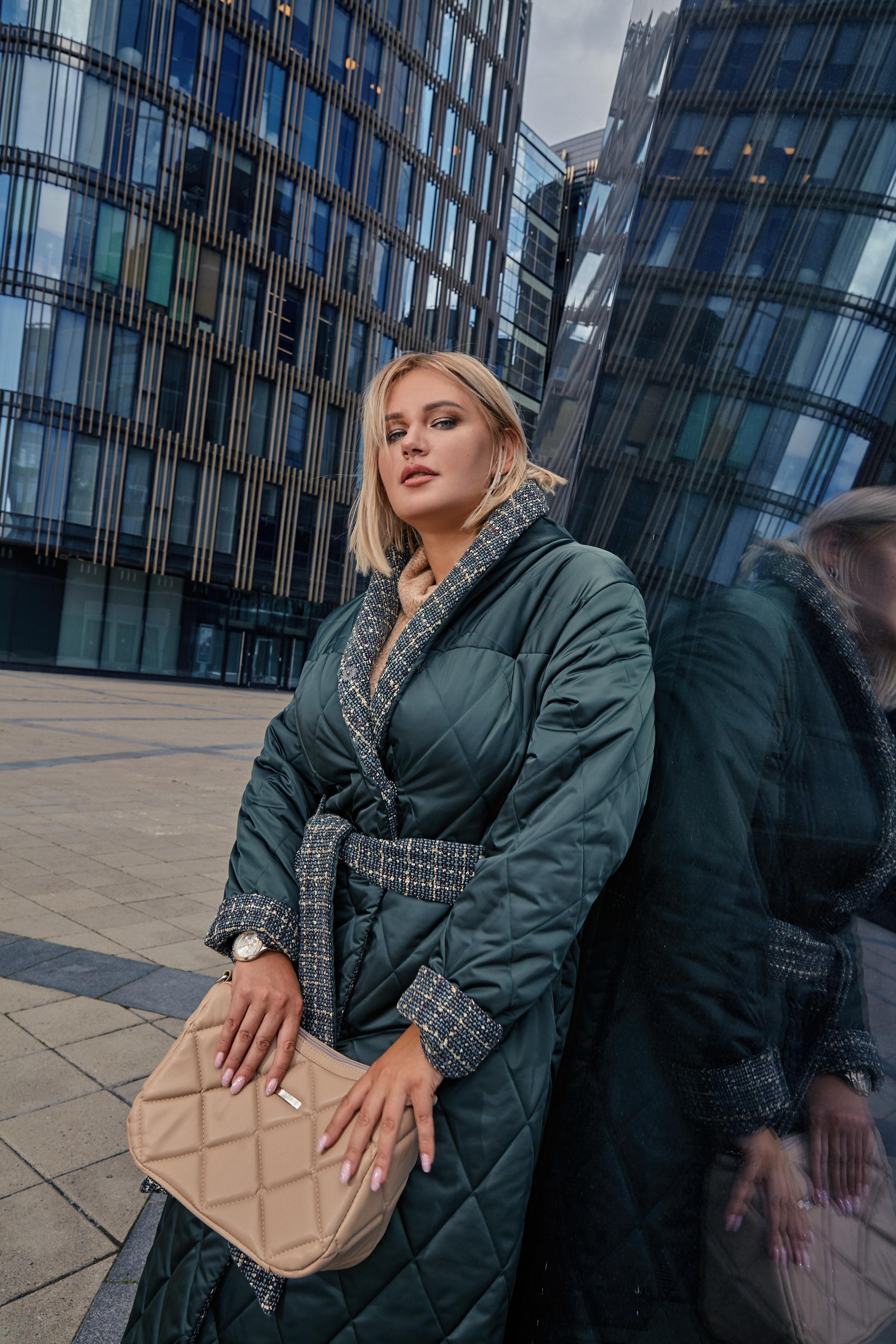 С.Жукова для Плюссайз зимней дежды Eva Graffova в зелёном пальто и бежевой сумкой на фоне стеклянных зданий