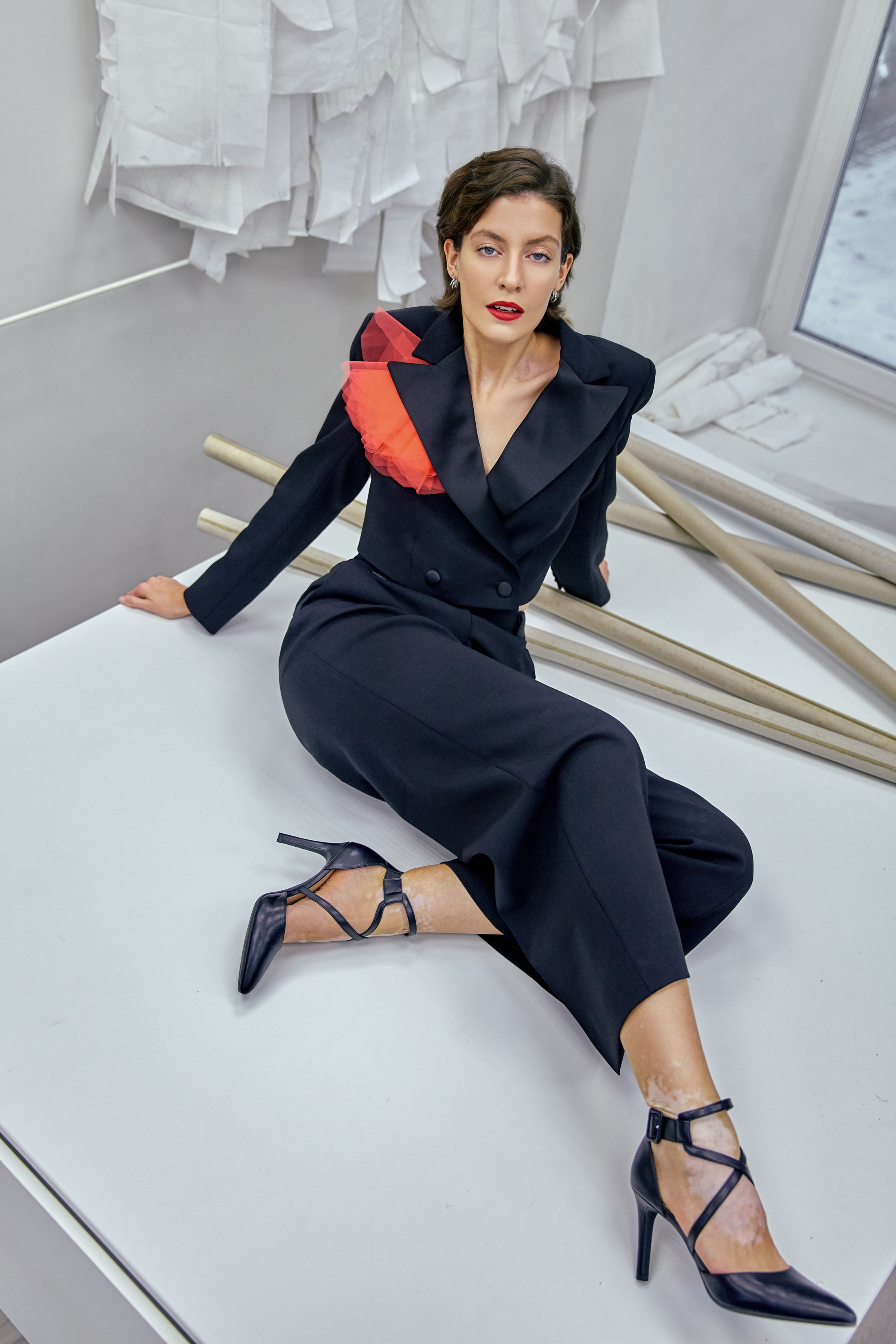 Модель с витилиго в черном дизайнерском костюме с красным маком Atelier star people сидит на закроечном столе