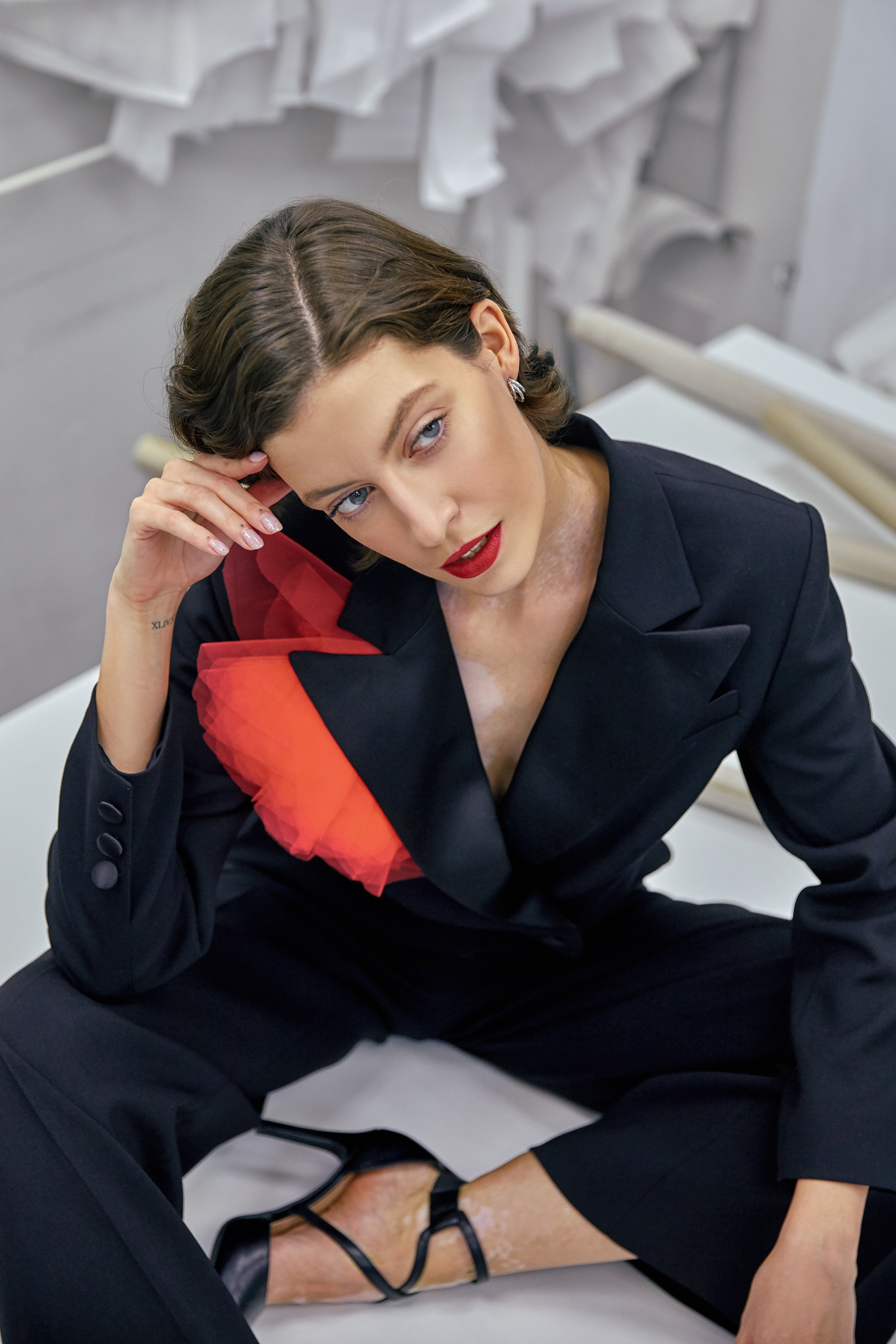 Девушка с витилиго в дизайнерском черном костюме с красным цветком сидит на закроечном столе