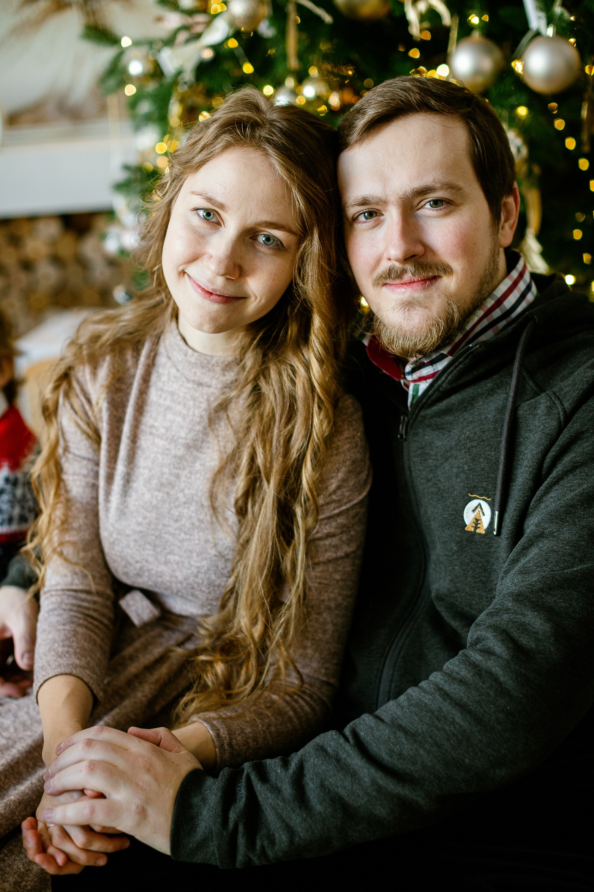 Семейная сидит на полу на фоне стоит елка и светится огоньками. Муж и жена смотрят в камеру.