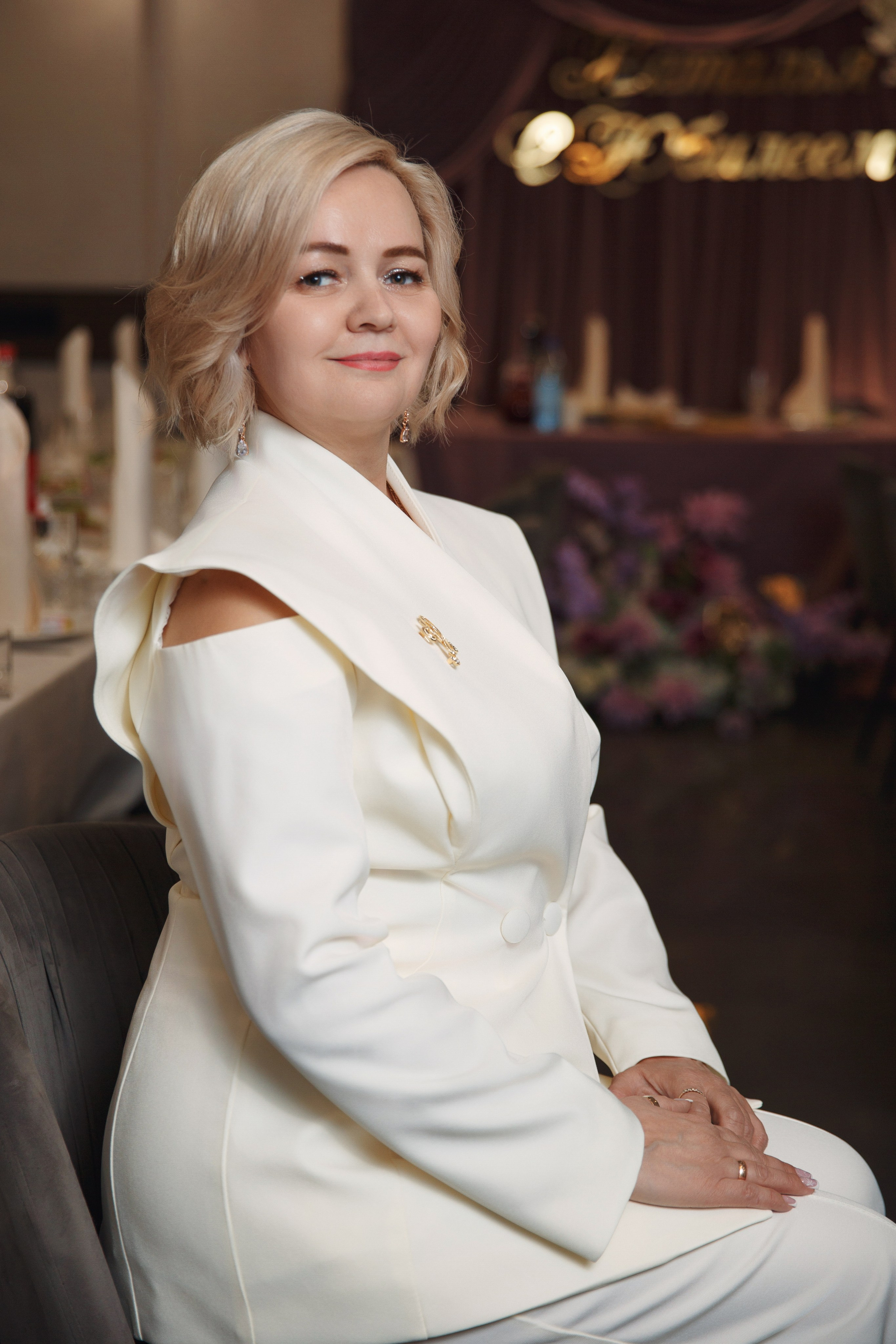 Именинница в белом праздничном костюме позирует в ресторане.