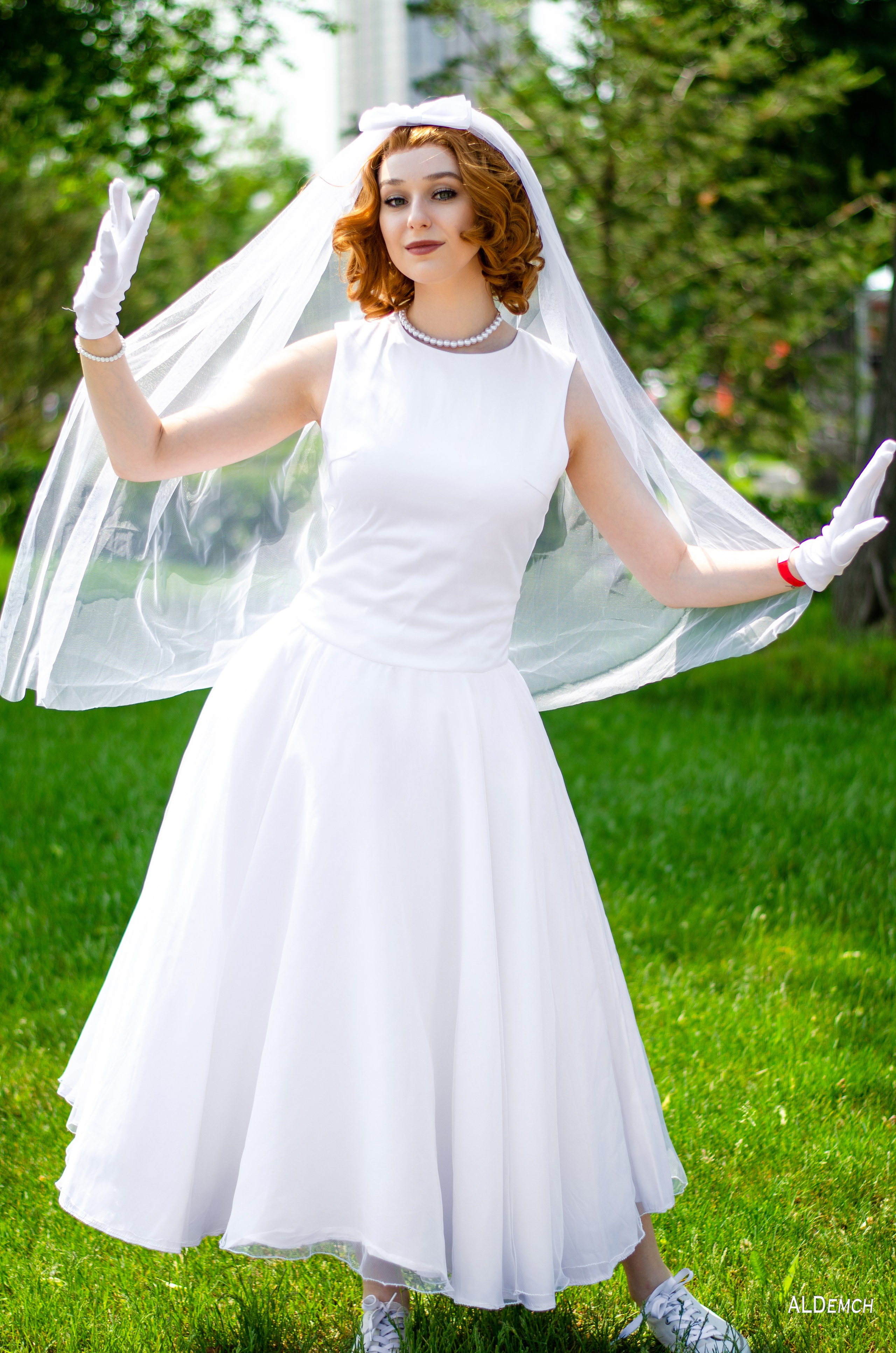 На фотографии изображена женщина в белом свадебном платье с фатой. У нее рыжие волосы, уложенные в локоны. Она стоит на зеленой траве на улице, на фоне деревьев. Женщина одета в белые перчатки и белые туфли. На шее у нее жемчужное ожерелье. Она улыбается и держит руки поднятыми в стороны. свадебное платье из осенней коллекции. Свадебное платье, Мантия.