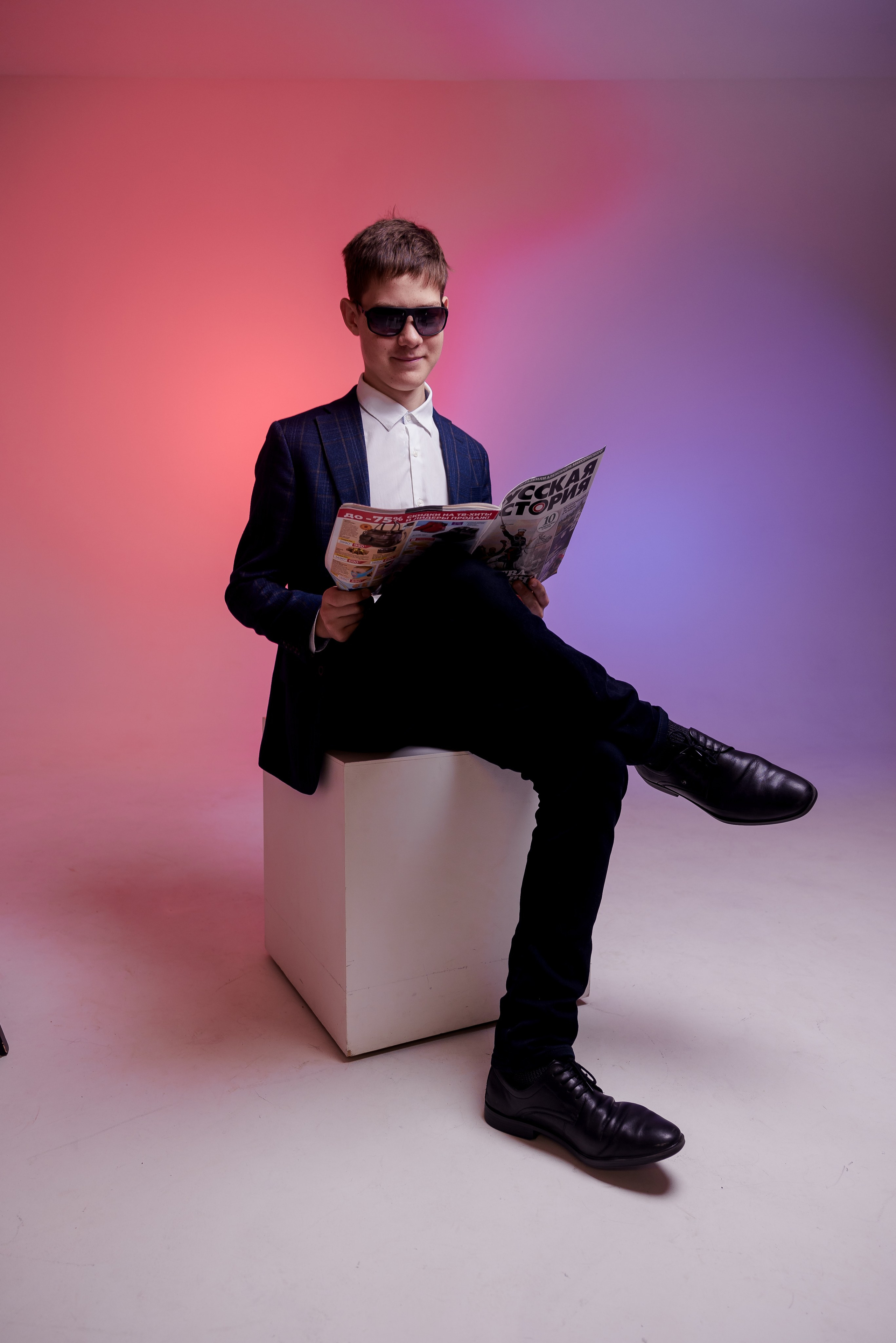 Молодой человек сидит на тумбе с газетой и в очках в фотостудии с цветным светом