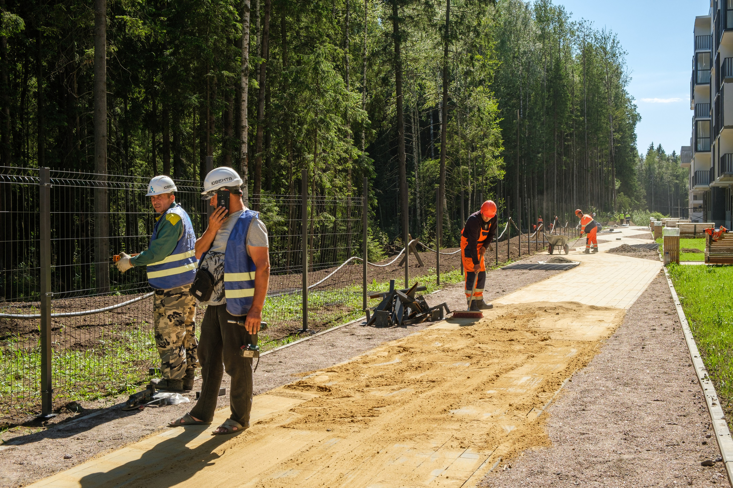 На фотографии изображен строительный объект, где работают люди в спецодежде. Они занимаются укладкой тротуарной плитки, а так же возведением забора.