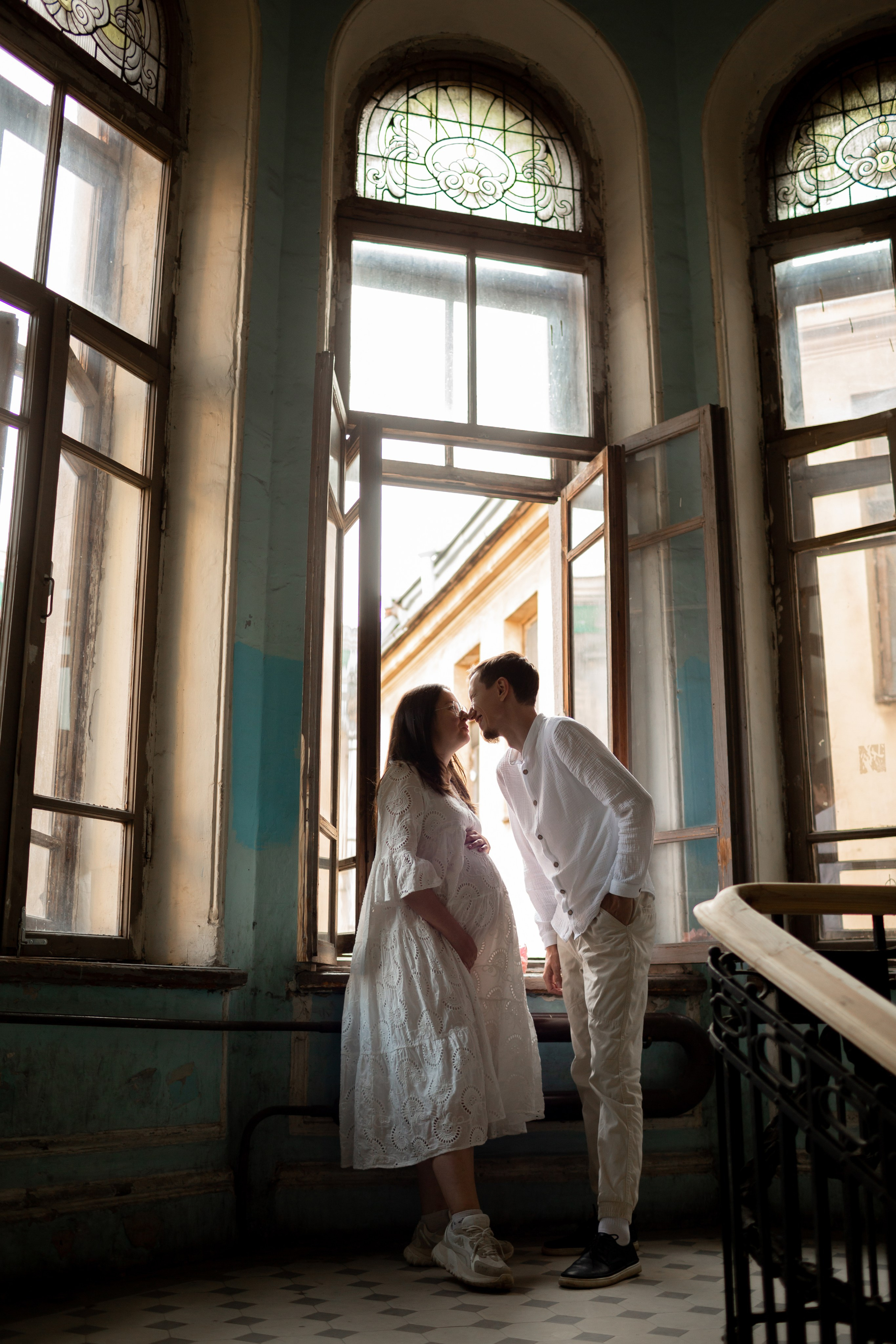 Беременная в белом платье стоит у окна исторической парадной.