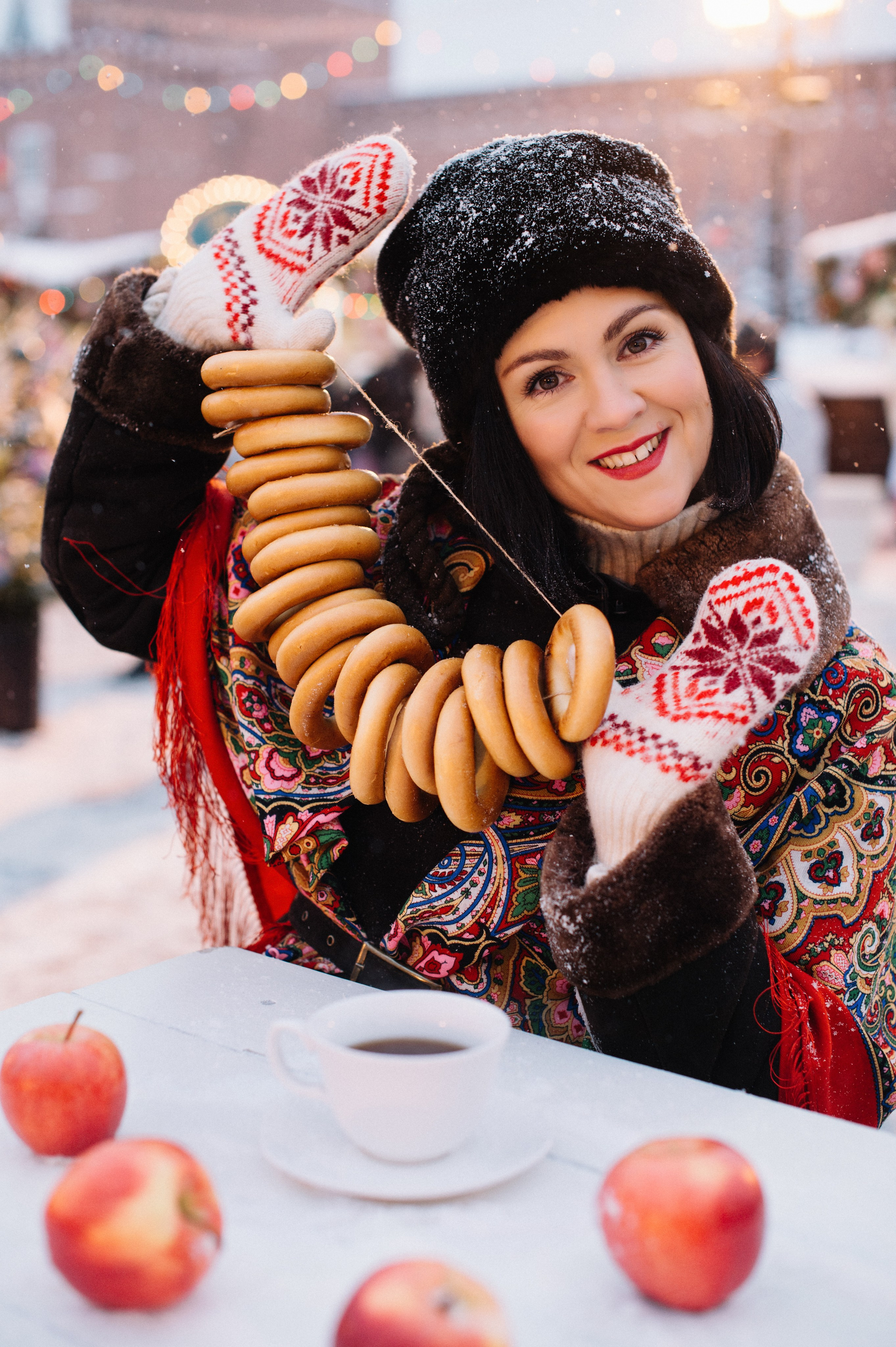 Девушка в красивом наряде в русском стиле с красным платком на плечах на Ярмарке на Красной площади зимой. Она держит в руках вязанку баранок и улыбается. На столе лежат красные яблоки и стоит чашка чая.