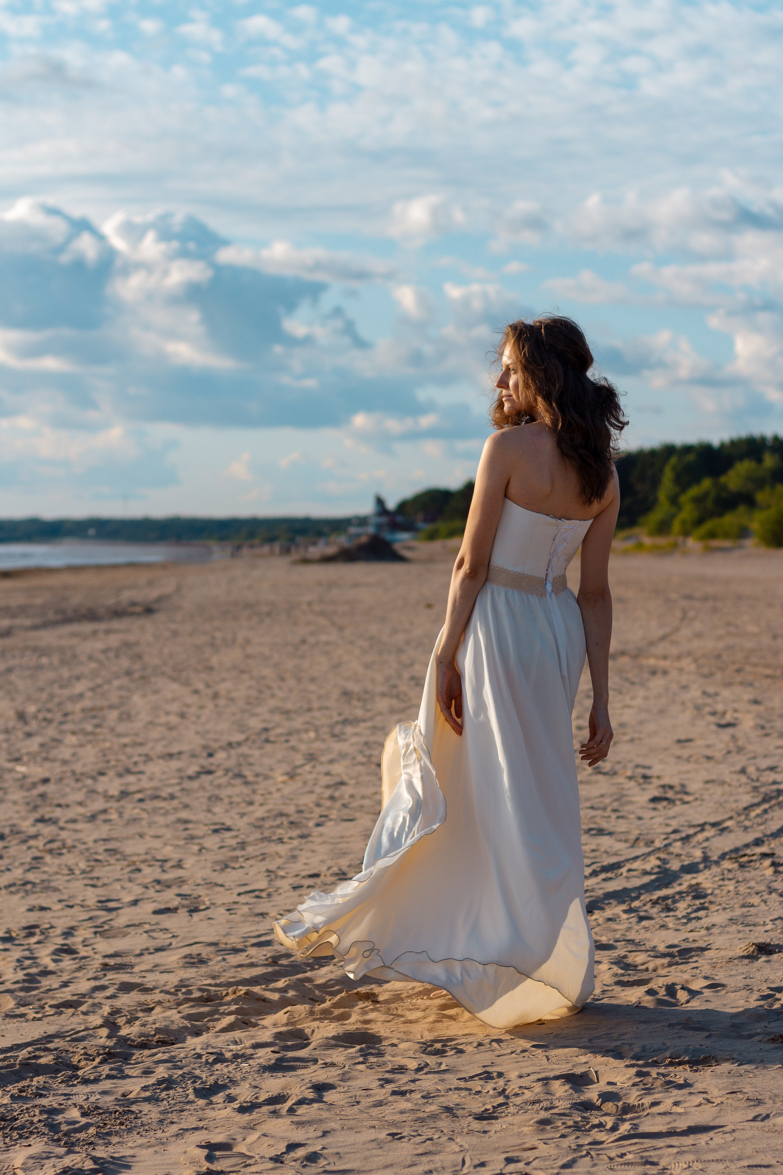 Фото со спины. Берег залива. Девушка со спины в открытом платье. Голубое небо с облаками. Воздушное развивающееся платье на ветру.