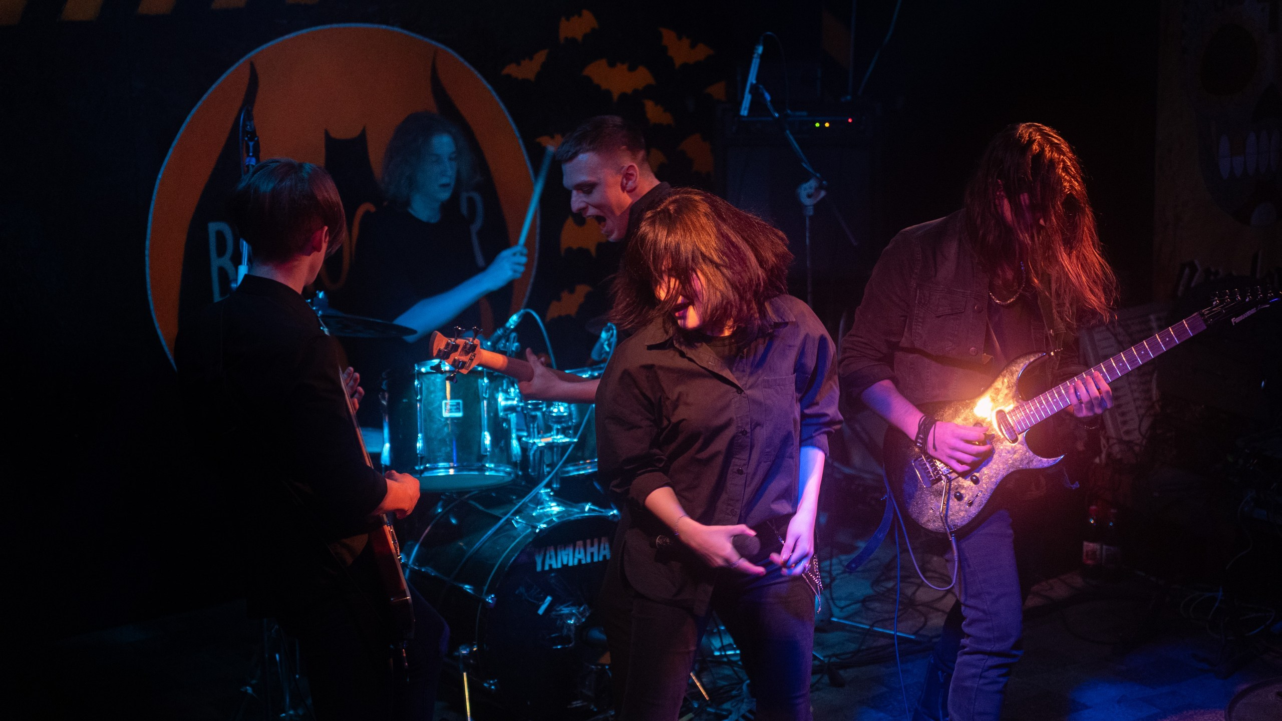 Это изображение группы, играющей в темной комнате. Группа состоит из барабанщика, гитариста, басиста и певца. Барабанщик находится сзади, за ударной установкой. Гитарист находится в левой части изображения и играет на красно-черной электрогитаре. Басист находится в правой части изображения и играет на черной бас-гитаре. Певец находится впереди, в центре изображения, и поет в микрофон. Все участники группы смотрят на певца. Фон темный, на группу светит прожектор.