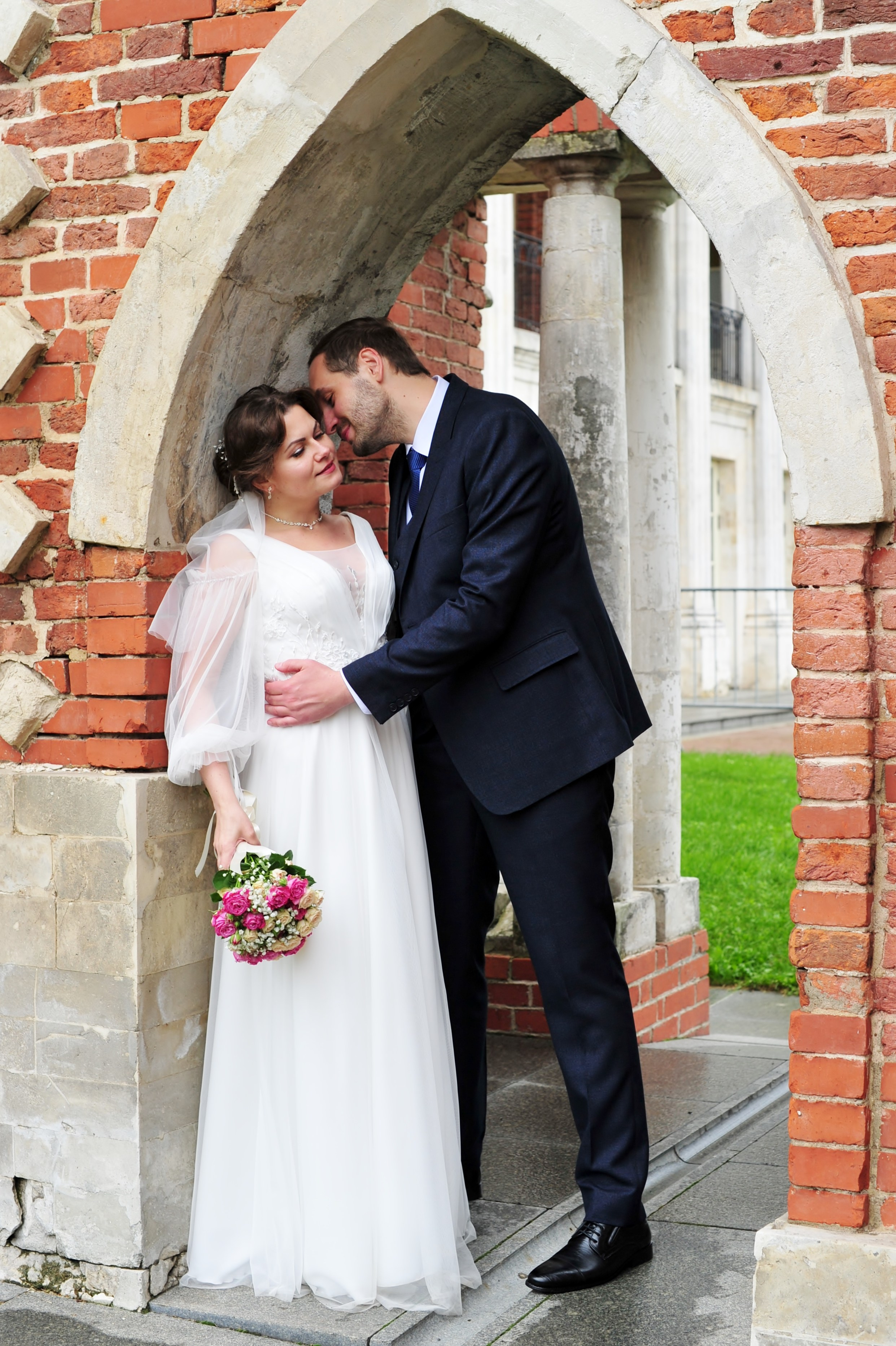Свадьба в Царицыно пасмурным летним днем, молодожены обнимаются в арке из красного кирпича