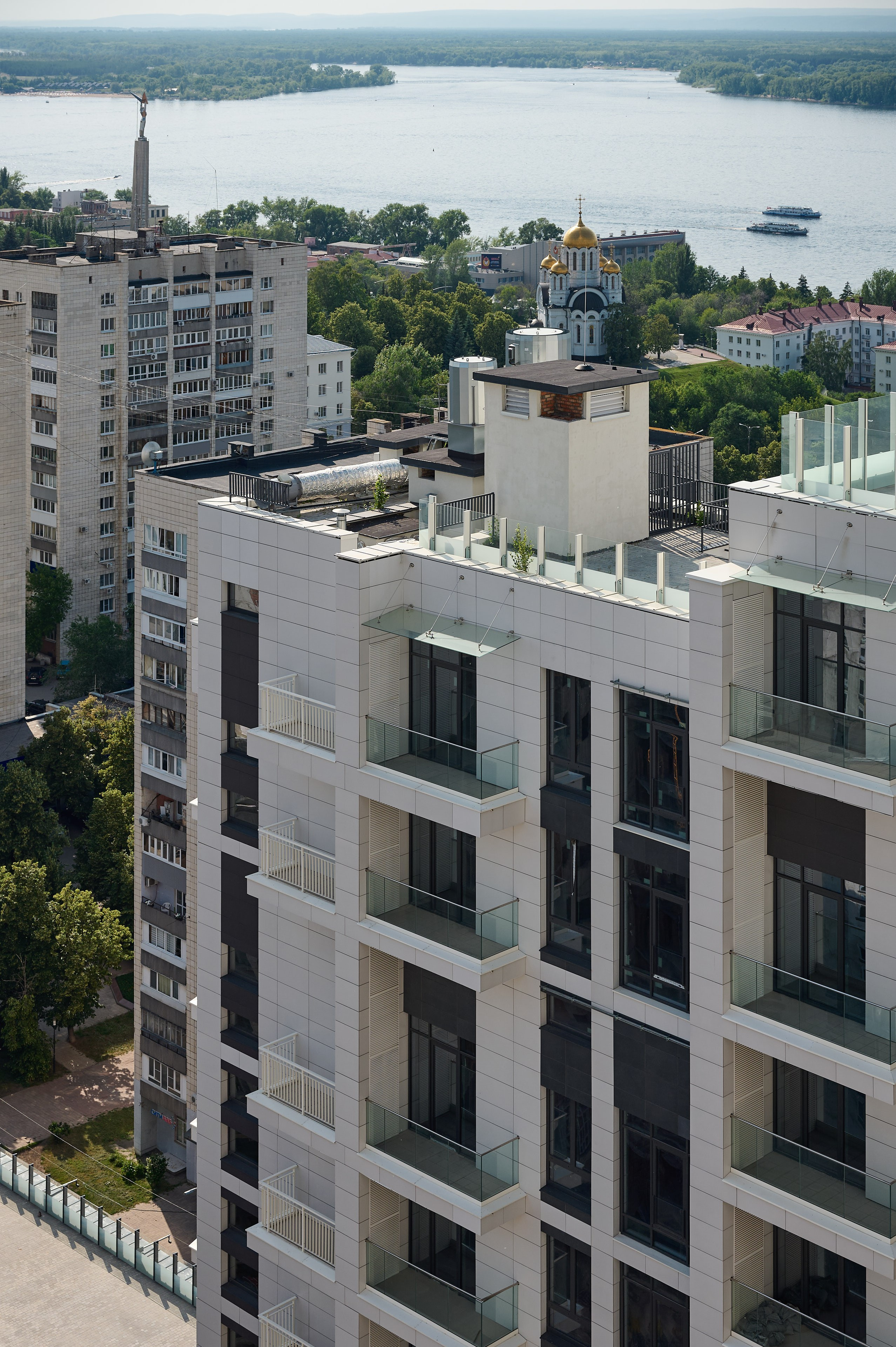 Вид на светлый жилой дом с современным фасадом на фоне городской застройки и широкой реки; в кадре также церковь с золотым куполом