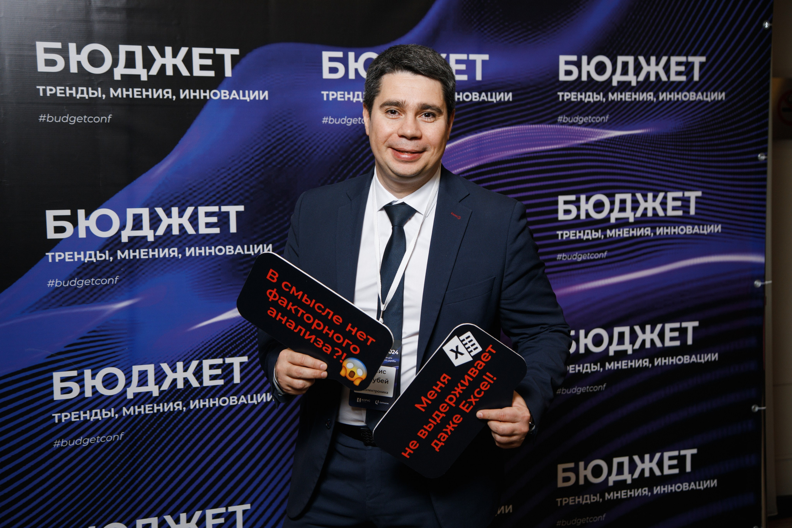 Мужчина в деловом костюме позирует в фотозоне конференции «Бюджет» с забавными табличками про Excel и факторный анализ на фоне синего брендированного стенда