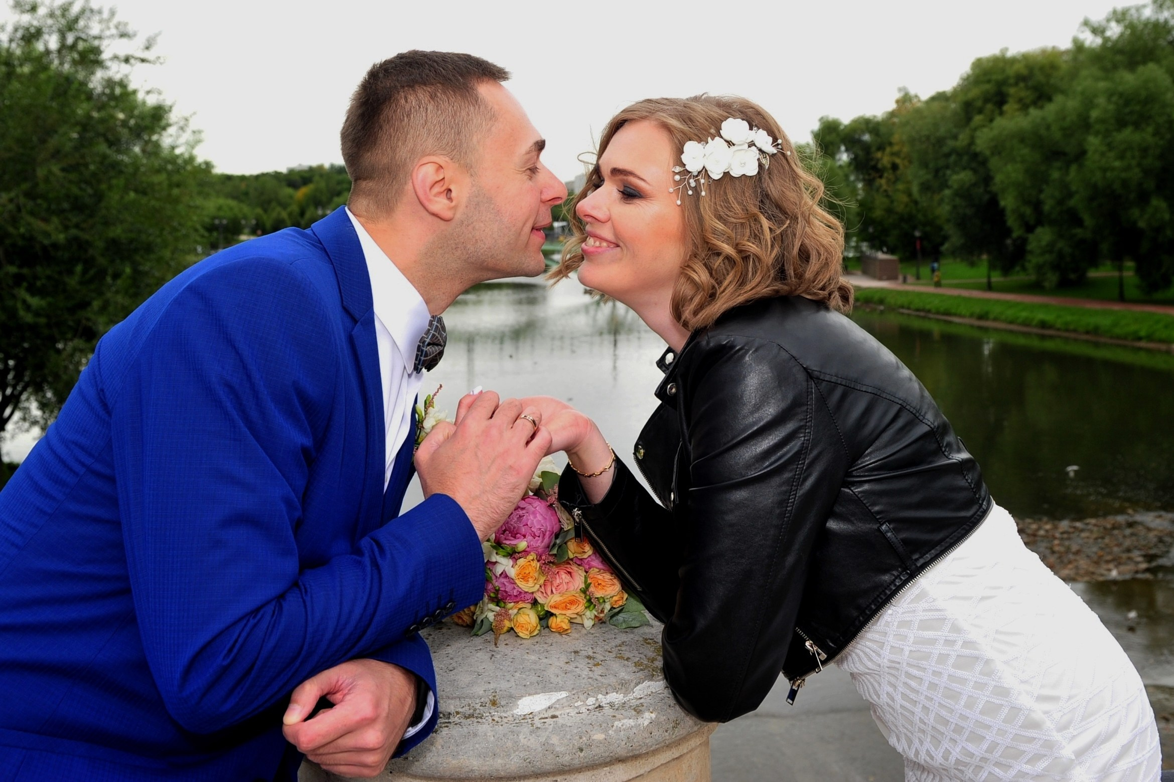 Свадьба в Царицыно хмурым прохладным летним днём, молодожены стоят на каменном мосту на фоне воды и держатся на руки, между ними лежит букет