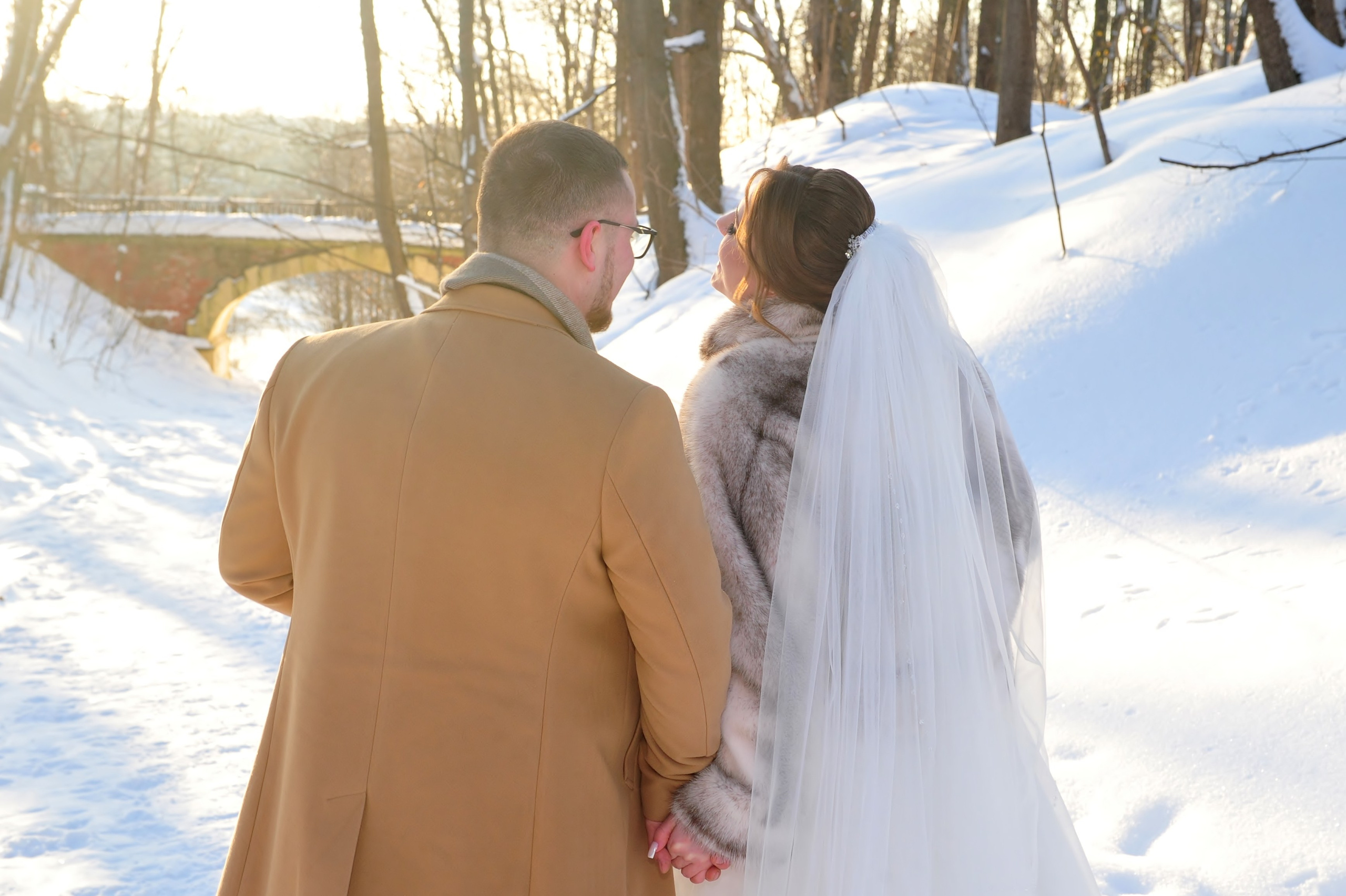 Зимняя свадьба в усадьбе Суханово, молодожены гуляют и держатся за руки, вид со спины на фоне каменного моста и арки