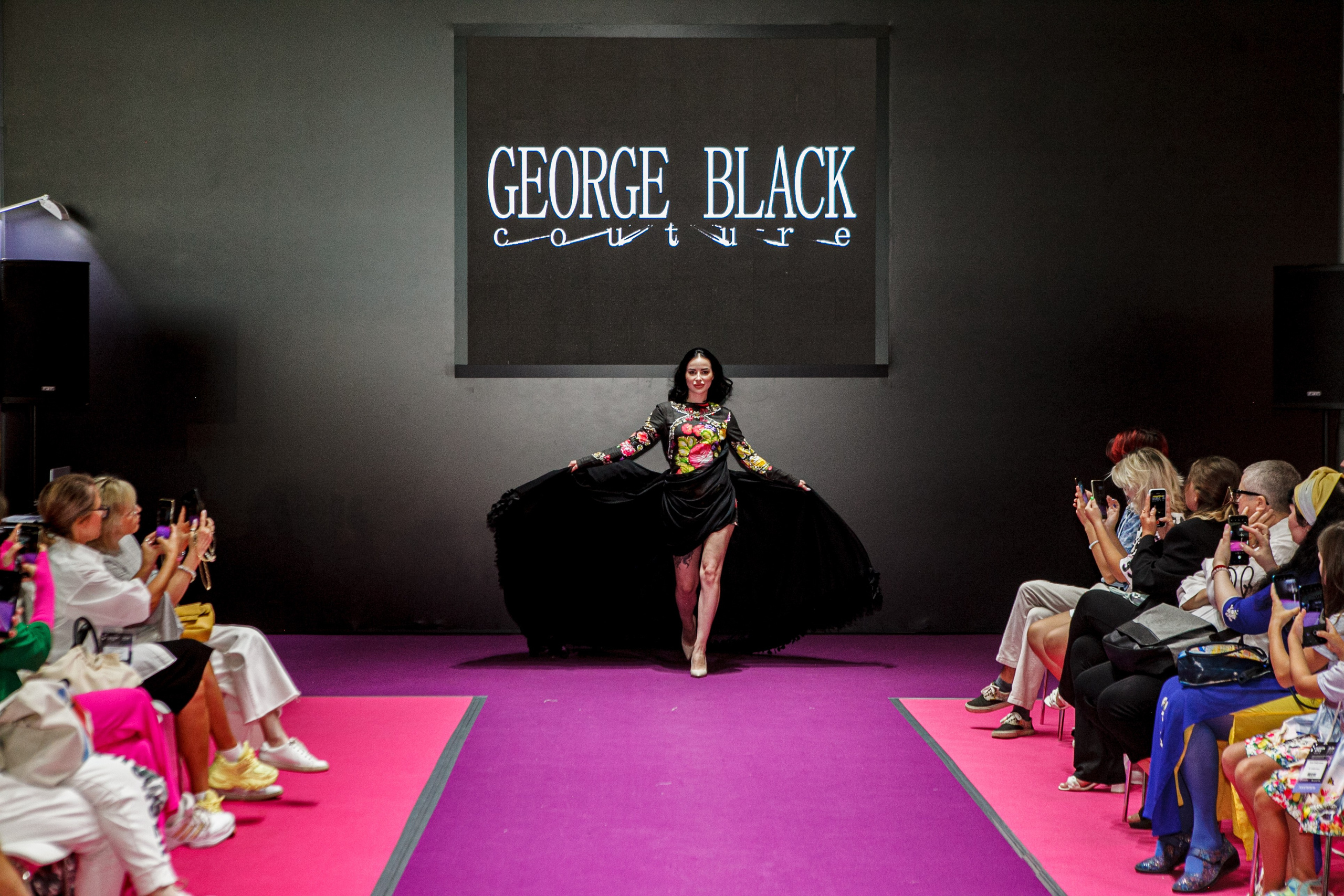  "Модели в ярких нарядах проходят по подиуму во время показа мод, освещенные яркими софитами и окруженные восторженными зрителями."