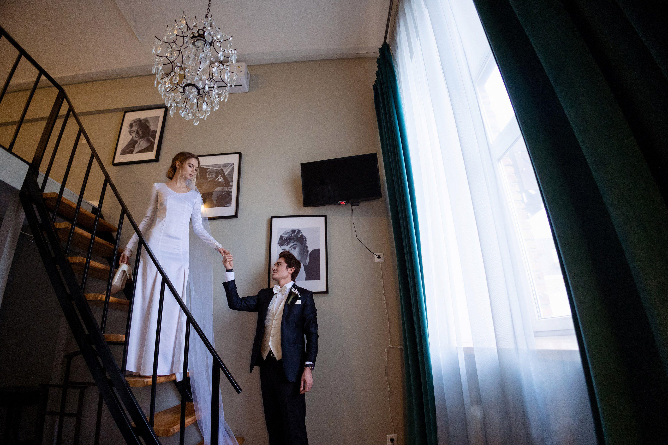 Жених помогает спускаться по лестнице не весте на фоне окна.