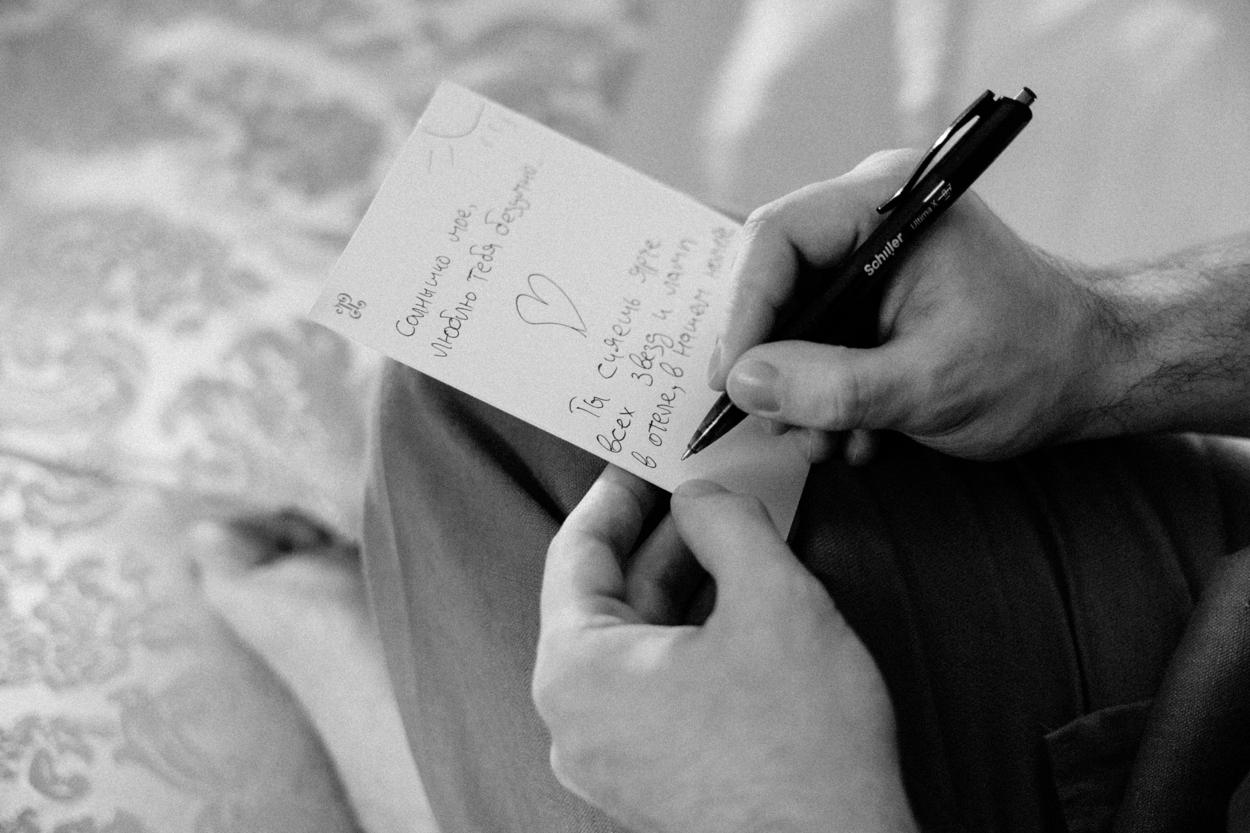 Черно-белое фото мужской руки, пишущей ручкой на открытке.