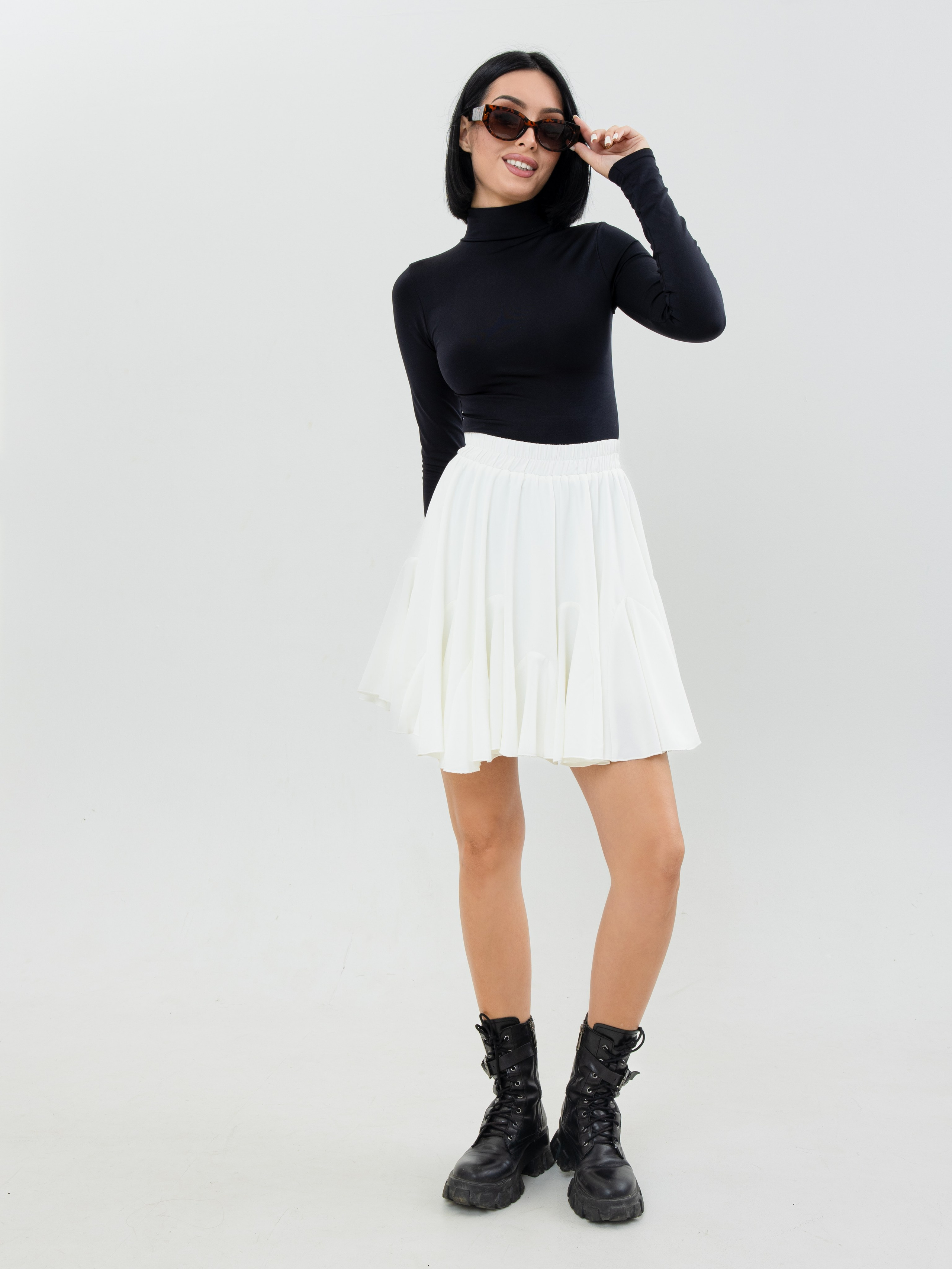 Девушка брюнетка в черной водолазке и белой юбке и в солнцезащитных очках стоит на белом фоне