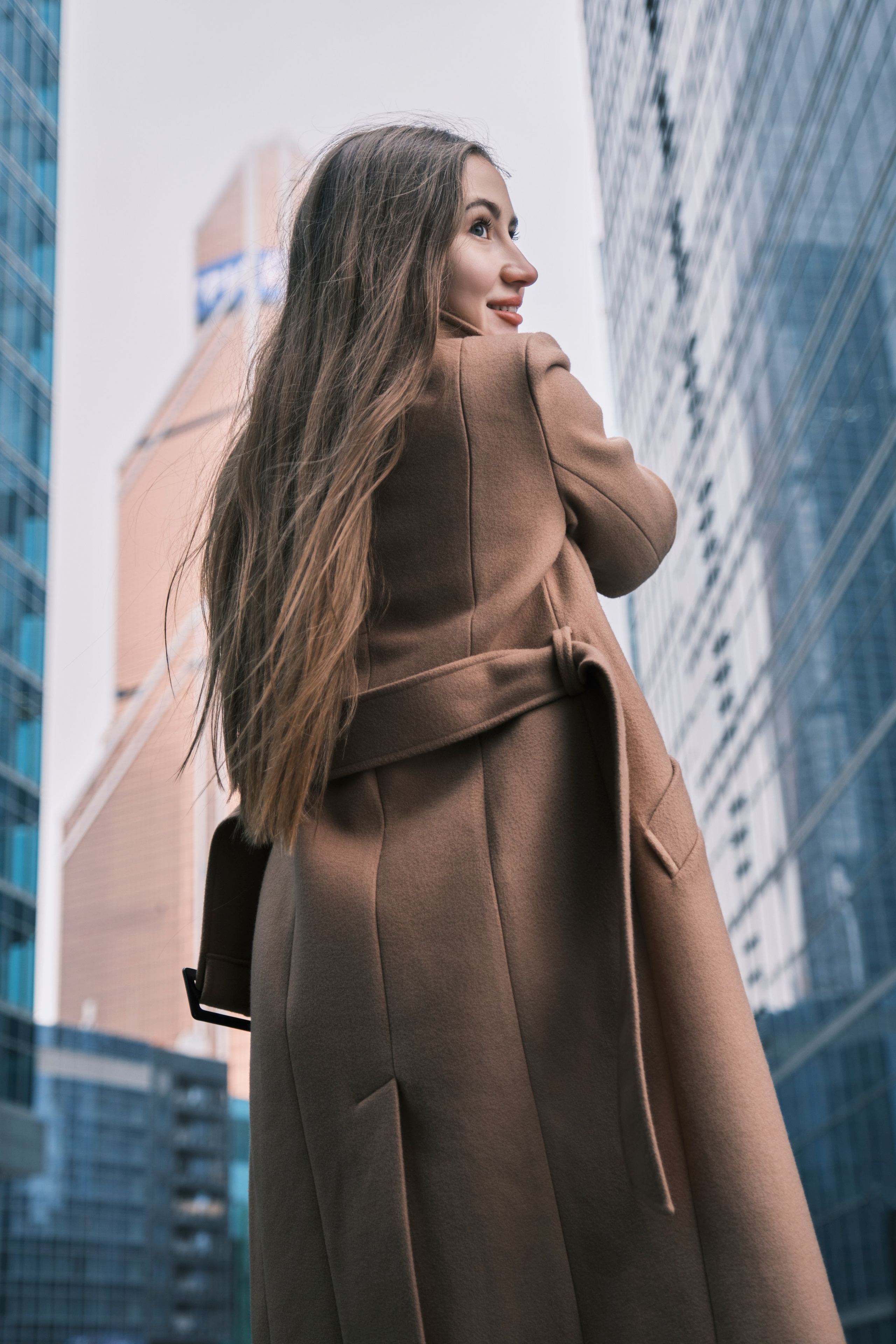 Девушка с длинными волосами в пальто стоит спиной и оборачивается в кадр на фоне башен Москва-Сити.