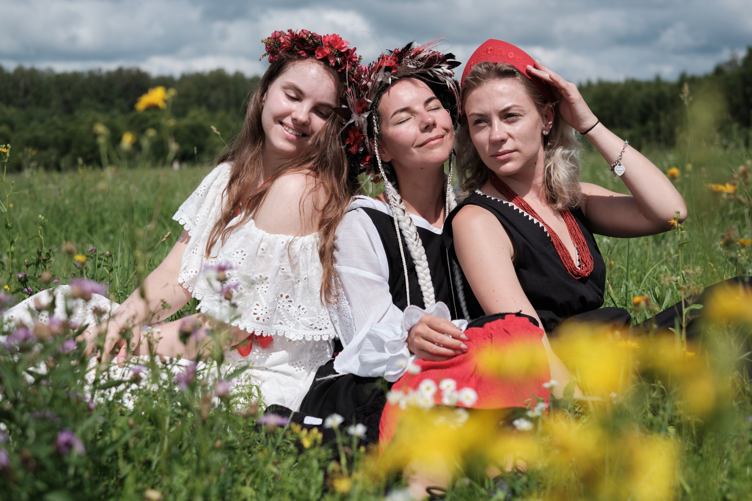 Три девушки в традиционных славянских нарядах и венках отдыхают на цветочном лугу.