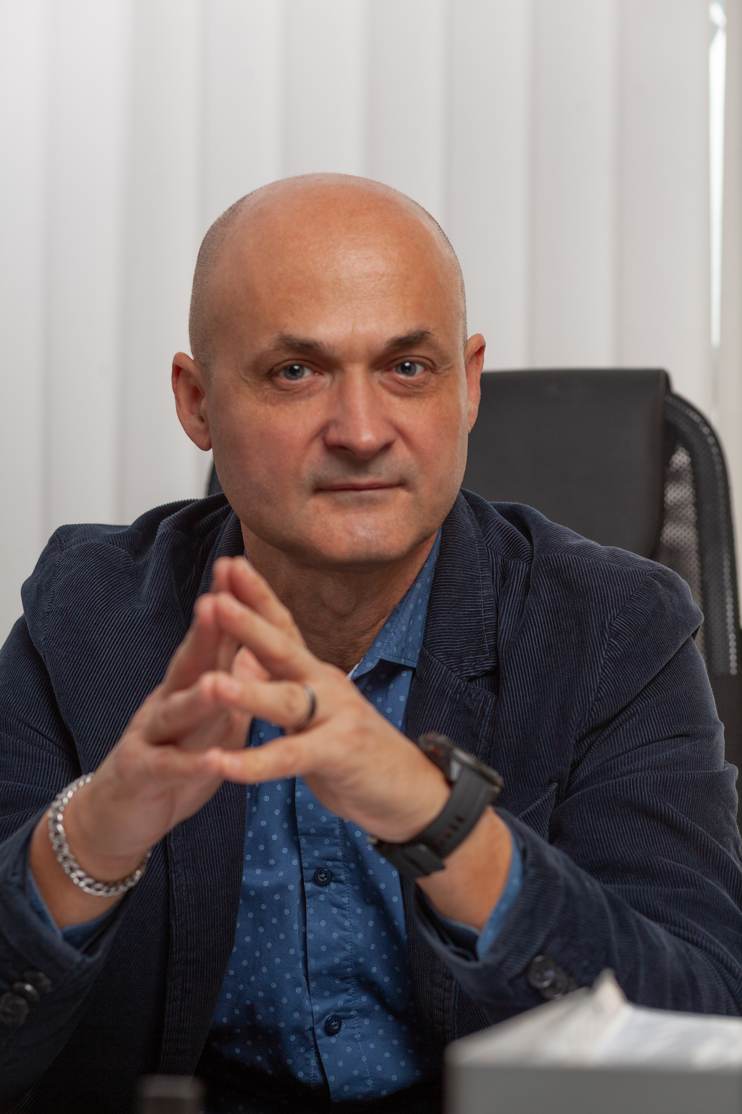 Лысый мужчина 50 лет сидит за столом сомкнув руки перед собой.