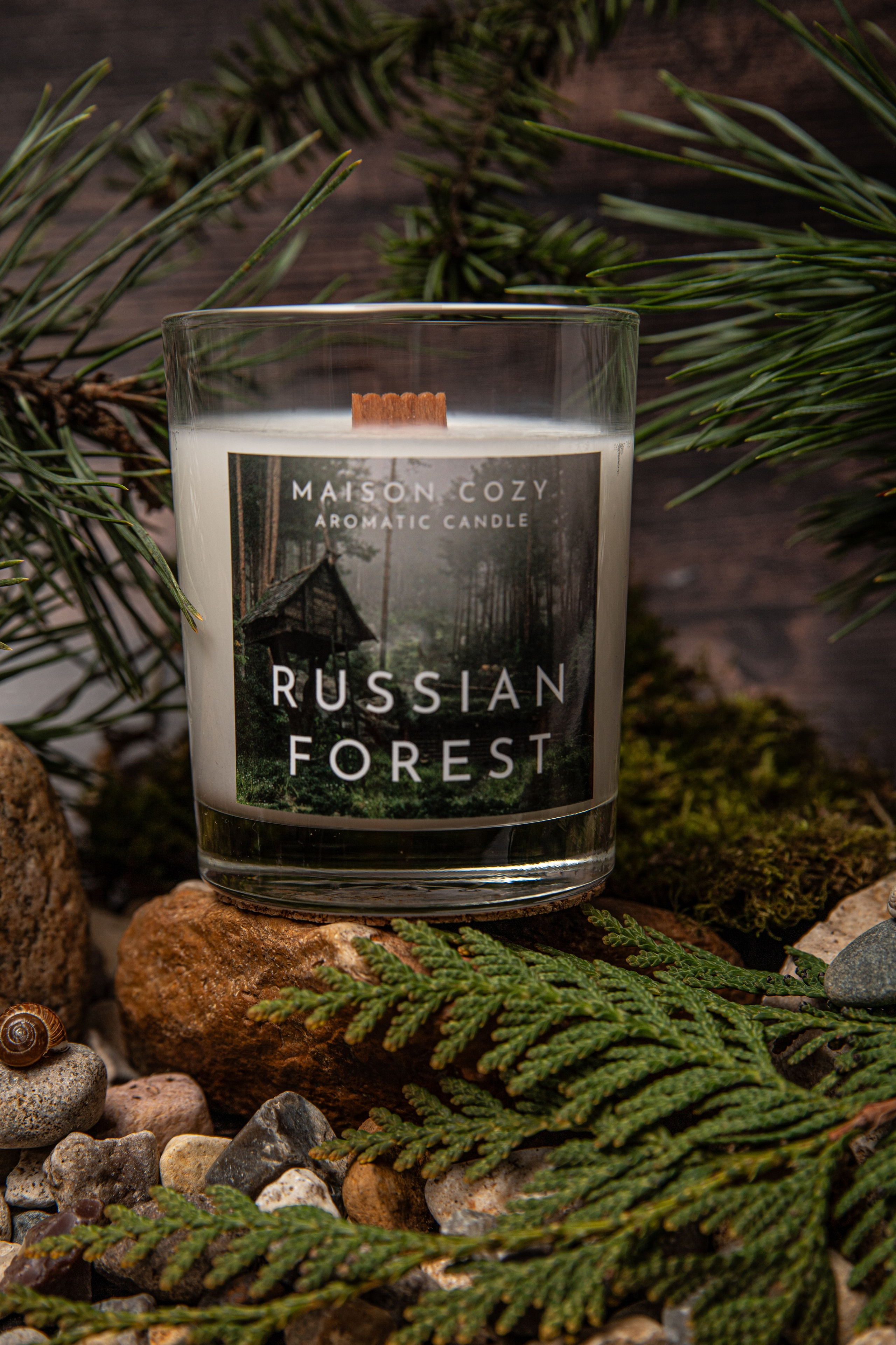 Ароматическая свеча “Русский лес” в прозрачном стеклянном стакане. Свеча окружена натуральной композицией: серыми камнями, ветками сосны и туи с зелёными хвоинками, а также мягким ковром из мха. Фон передаёт атмосферу хвойного леса — свежий, древесный аромат дополнен уютом природного убранства.