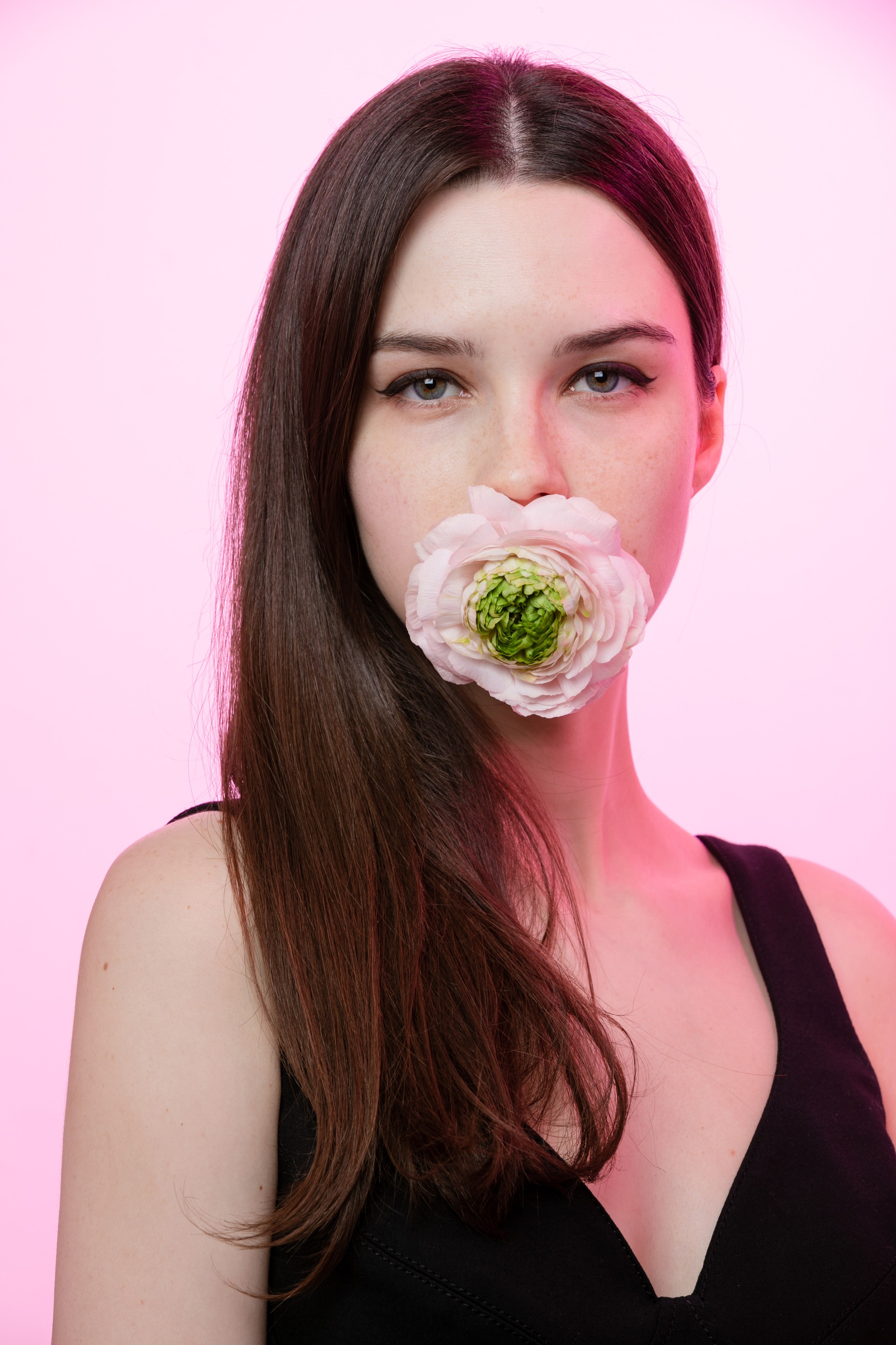 Девушка на розовом фоне держит во рту большой розовый цветок