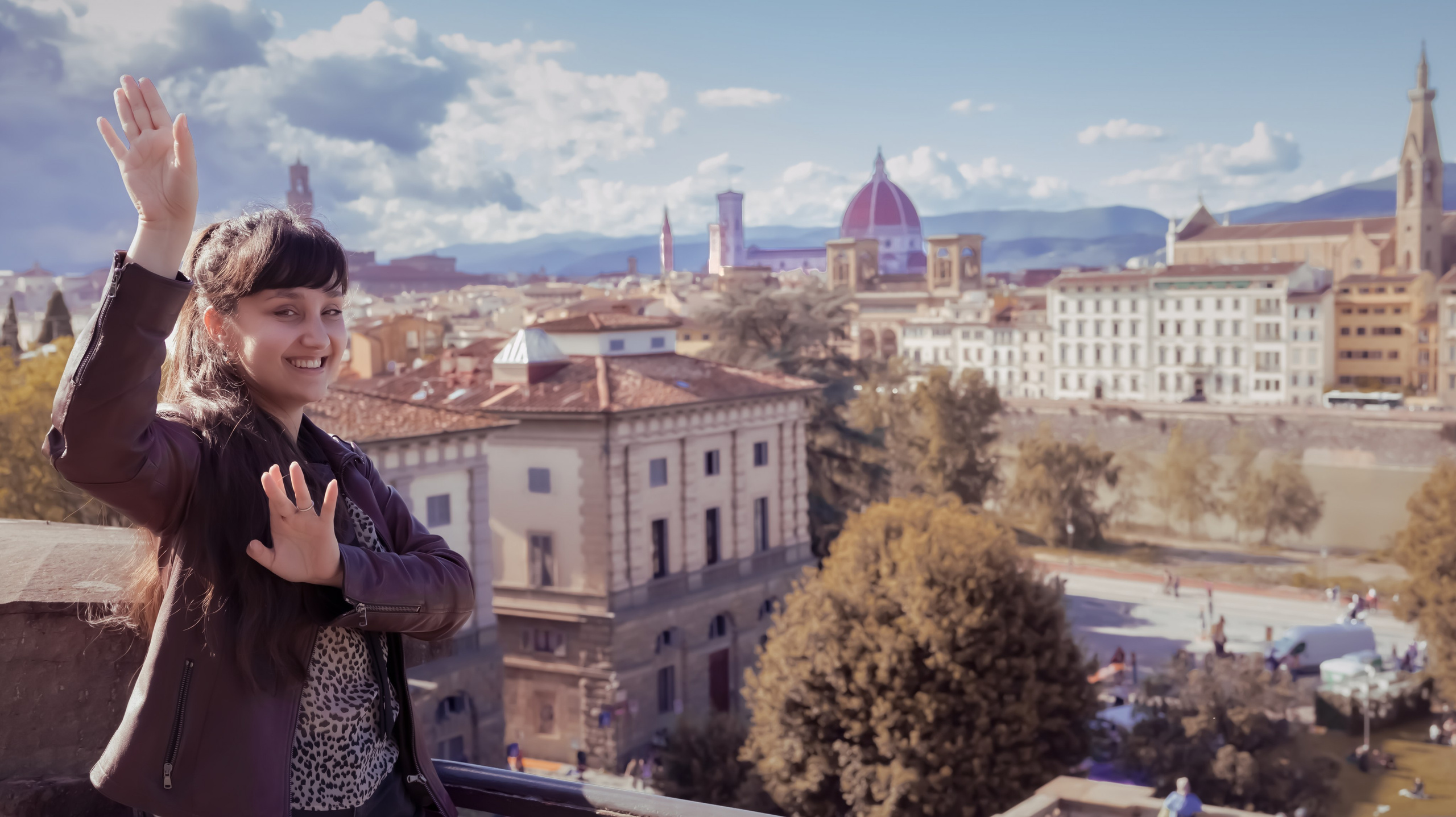Женщина в темной куртке и топе с леопардовым принтом широко улыбается и машет рукой, любуясь историческим пейзажем Флоренции с виднеющимся на заднем плане собором Дуомо.