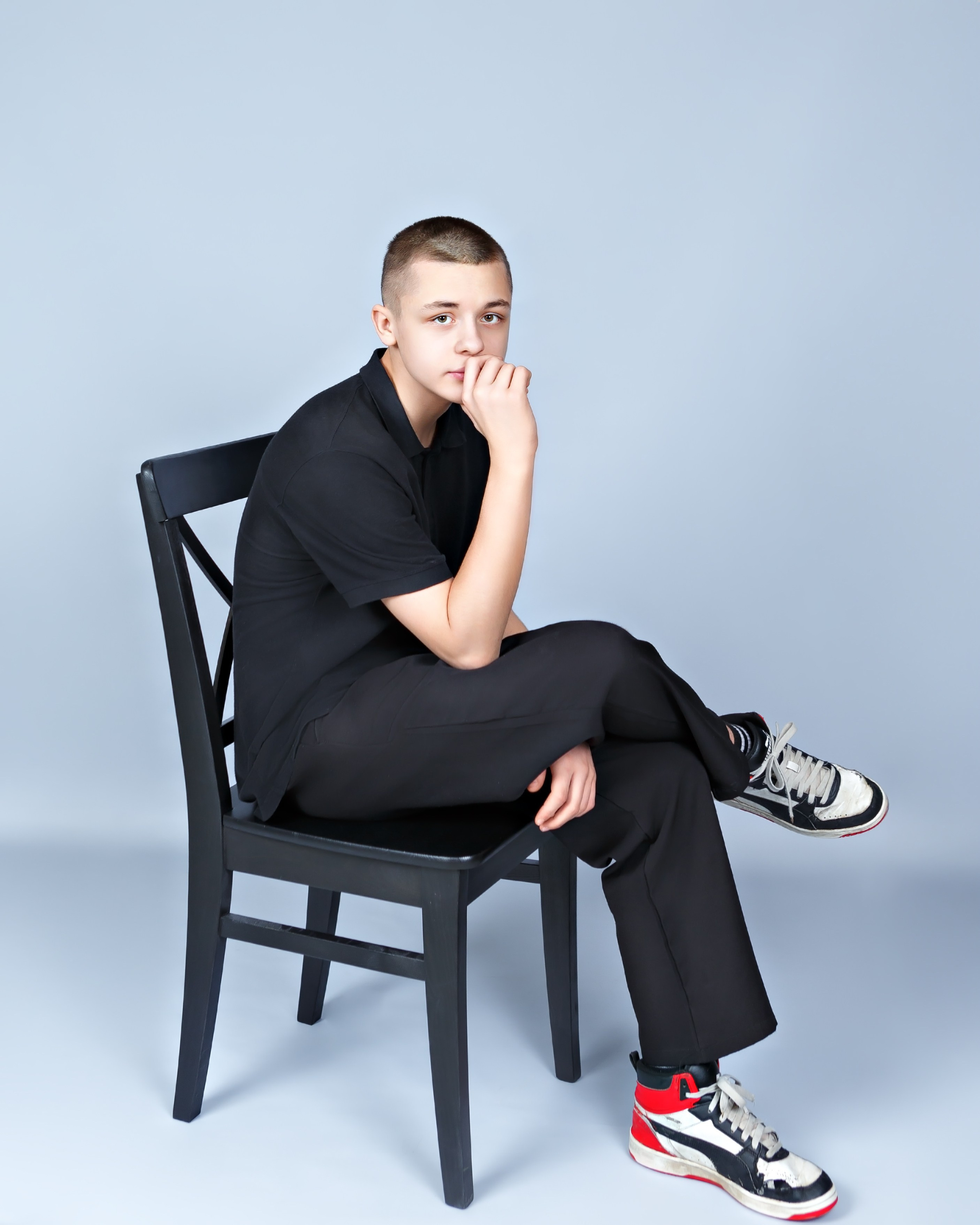 Портрет парня на стуле в черном костюме и кедах на сером фоне.